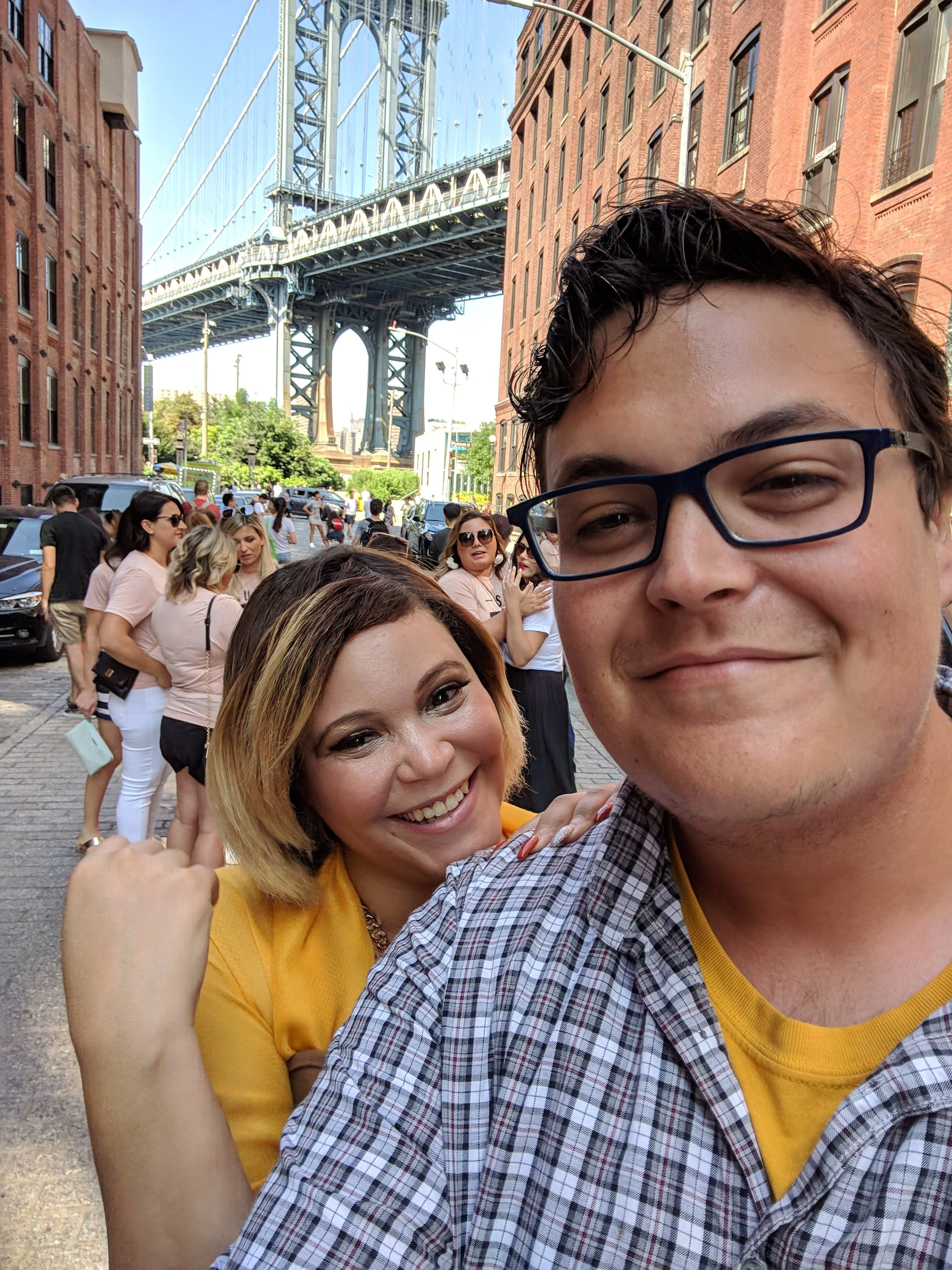 Two people taking a selfie on a city street with the Manhattan Bridge in the background. The woman is smiling, wearing a yellow top, and has shoulder-length blonde hair. The man is wearing glasses, a checkered shirt, and has short dark hair.