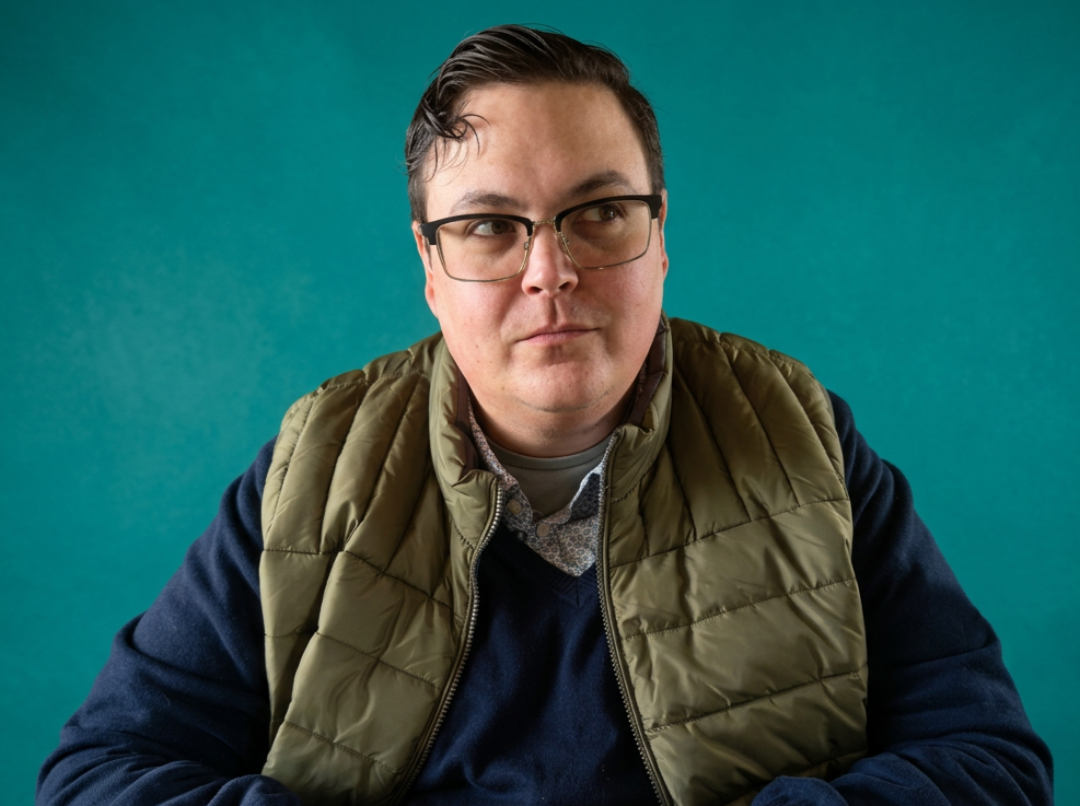 Frank, the co-founder, with glasses and dark hair, wearing a green puffy vest over a navy sweater, sitting against a teal background.