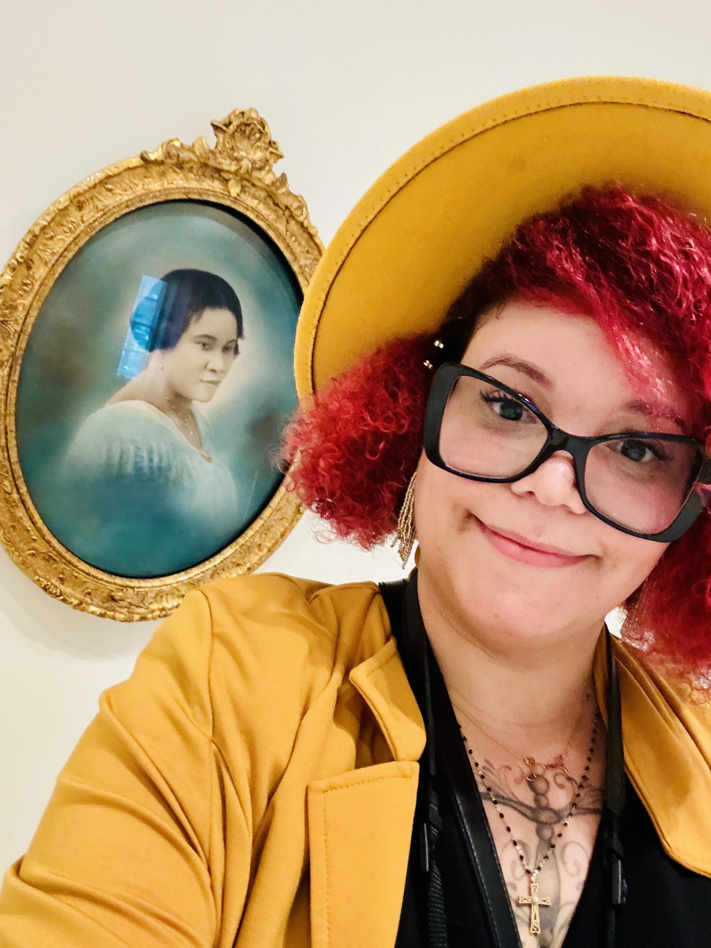 Nourich Founder, Briana McCrae Carr taking a selfie in front of a framed portrait. The woman has curly red hair, wears glasses, a large yellow hat, and a yellow jacket. The portrait of Madam C.J. Walker in the National Gallery is behind her.