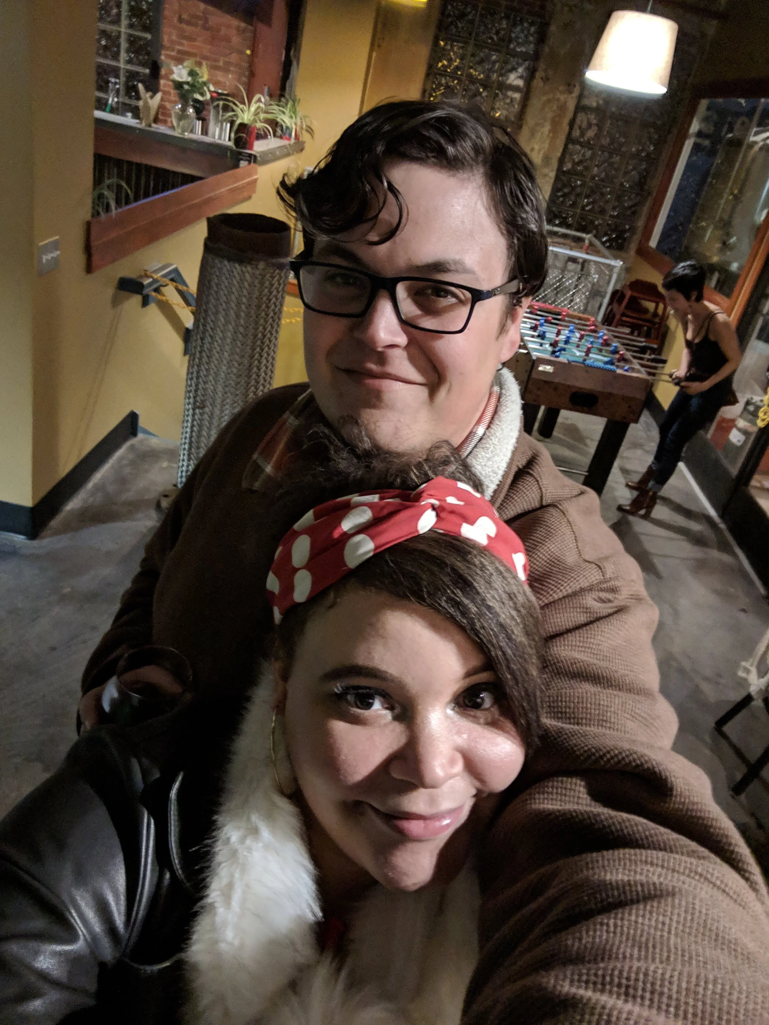 A man with glasses and dark hair smiling, captured in a selfie with a woman with dark hair and brown eyes, wearing a red polka dot headband. They are indoors with a foosball table and a woman playing in the background.