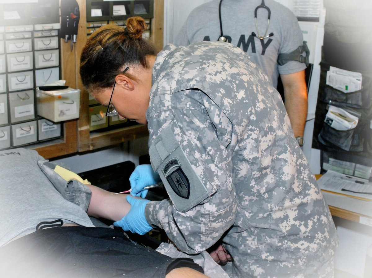 A female soldier in camouflage uniform drawing blood from a patient in a medical setting, with medical supplies and a person wearing a gray T-shirt with 'ARMY' printed on it in the background.
