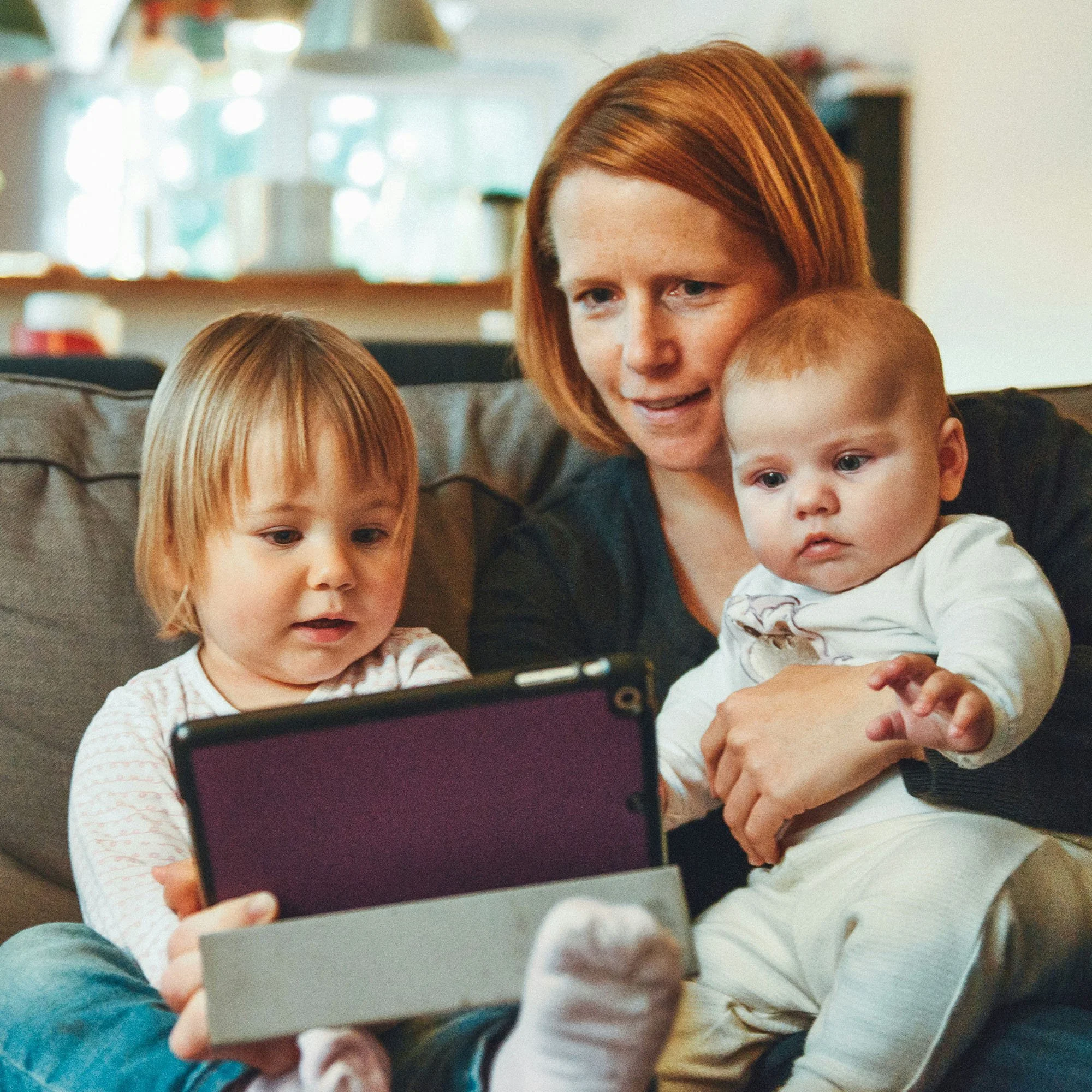 A woman with two young children looking at a tablet together on a couch in a cozy living room.