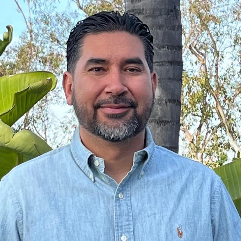 A middle-aged man with dark hair and a beard, wearing a light blue button-up shirt, standing outdoors in front of a tree and tropical plants.