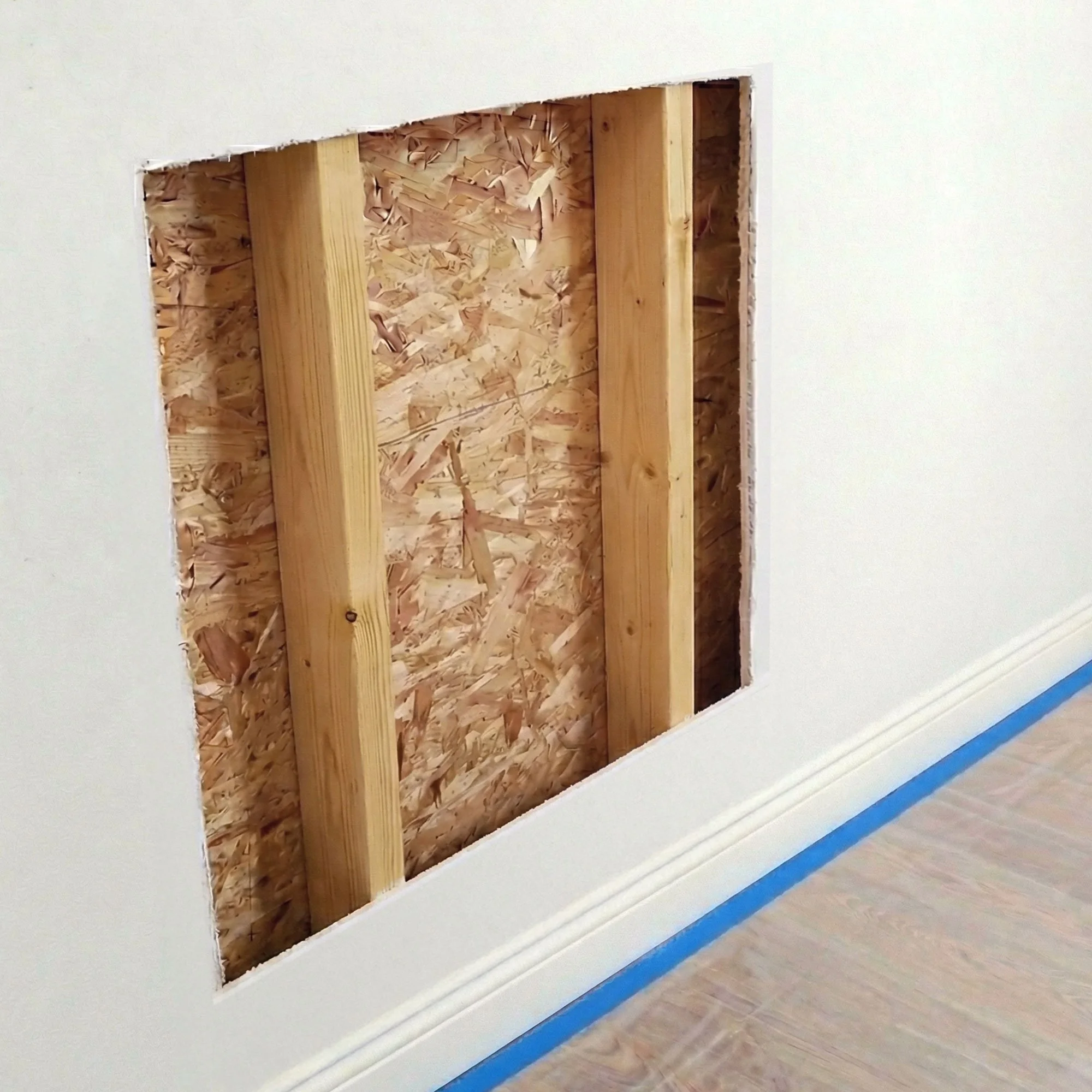 Opening in a wall showing exposed wood studs and oriented strand board subfloor, with painter's tape along the baseboard.