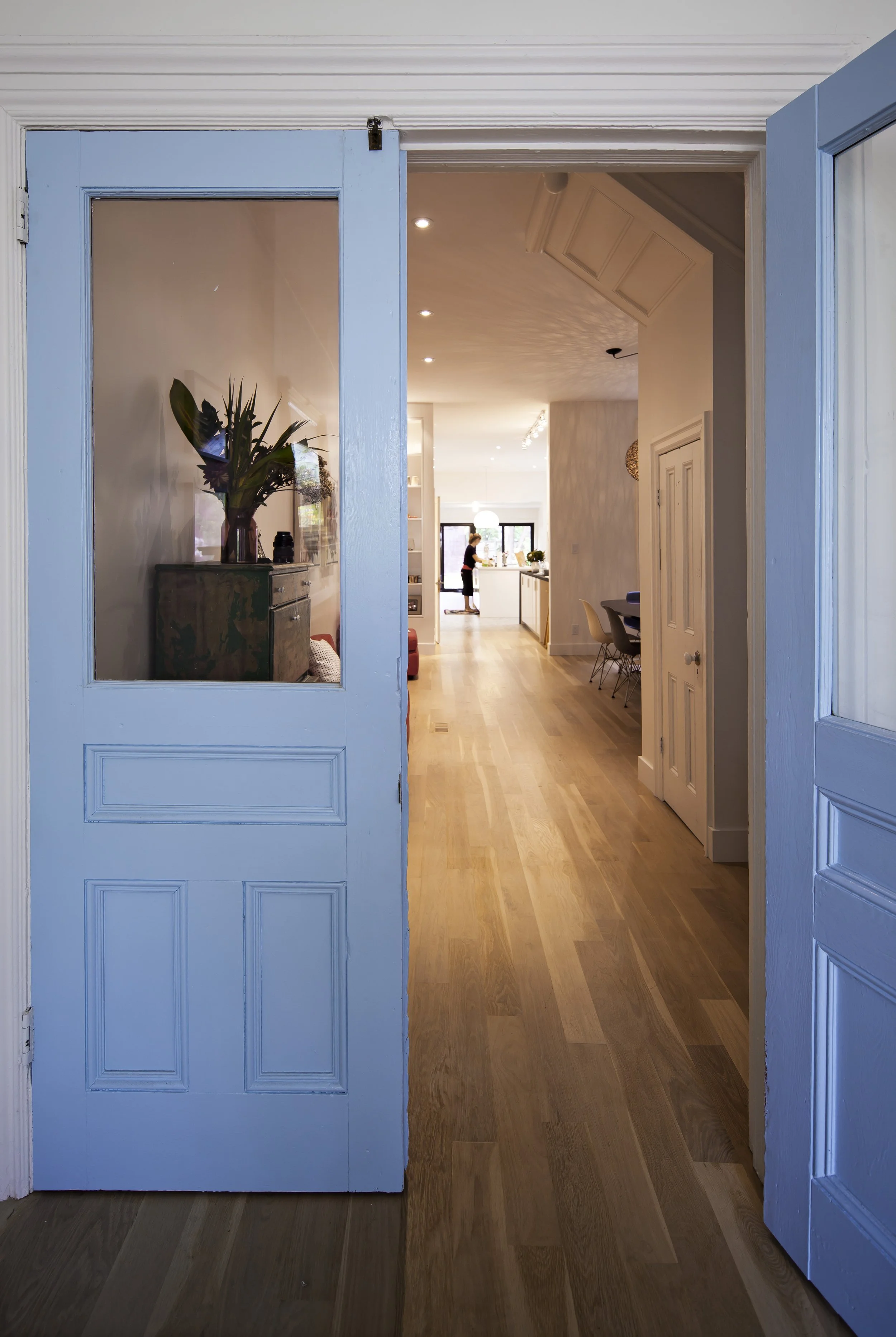 View through open blue double doors into a bright kitchen and dining area with wooden floors and a person standing inside.