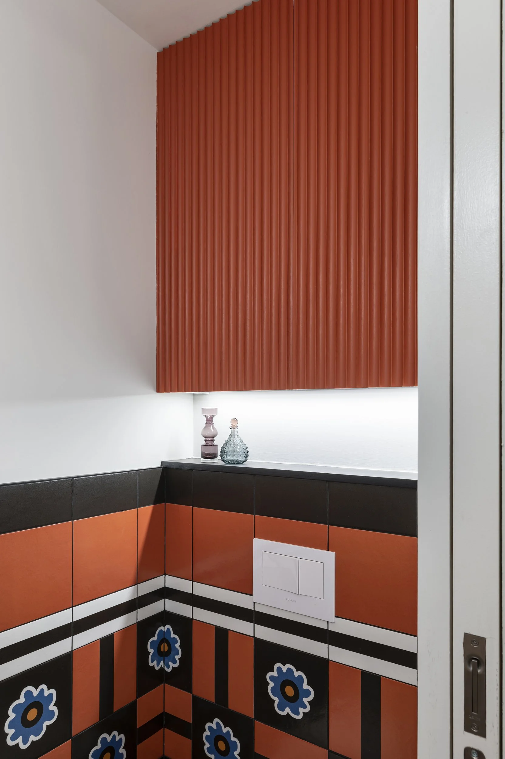 Interior corner of a bathroom with orange, black, and floral patterned tiles, a white toilet flush plate, and decorative bottles on a shelf, with a large orange textured cabinet on the wall.