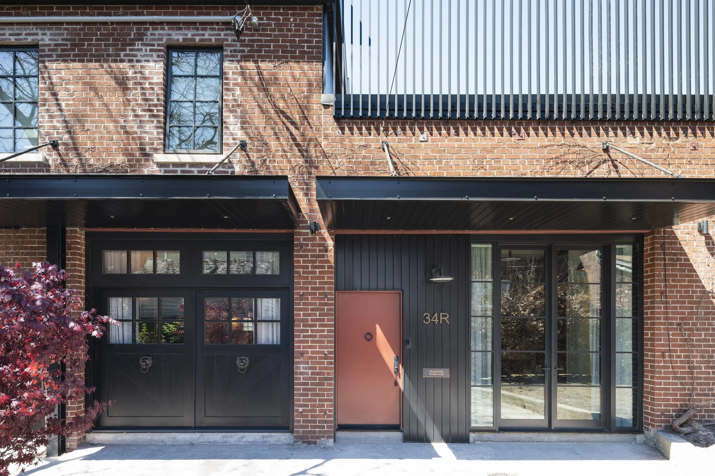 Modern townhouse with brick facade, black garage doors, a pink front door, the address 34R, and large glass windows reflecting trees.