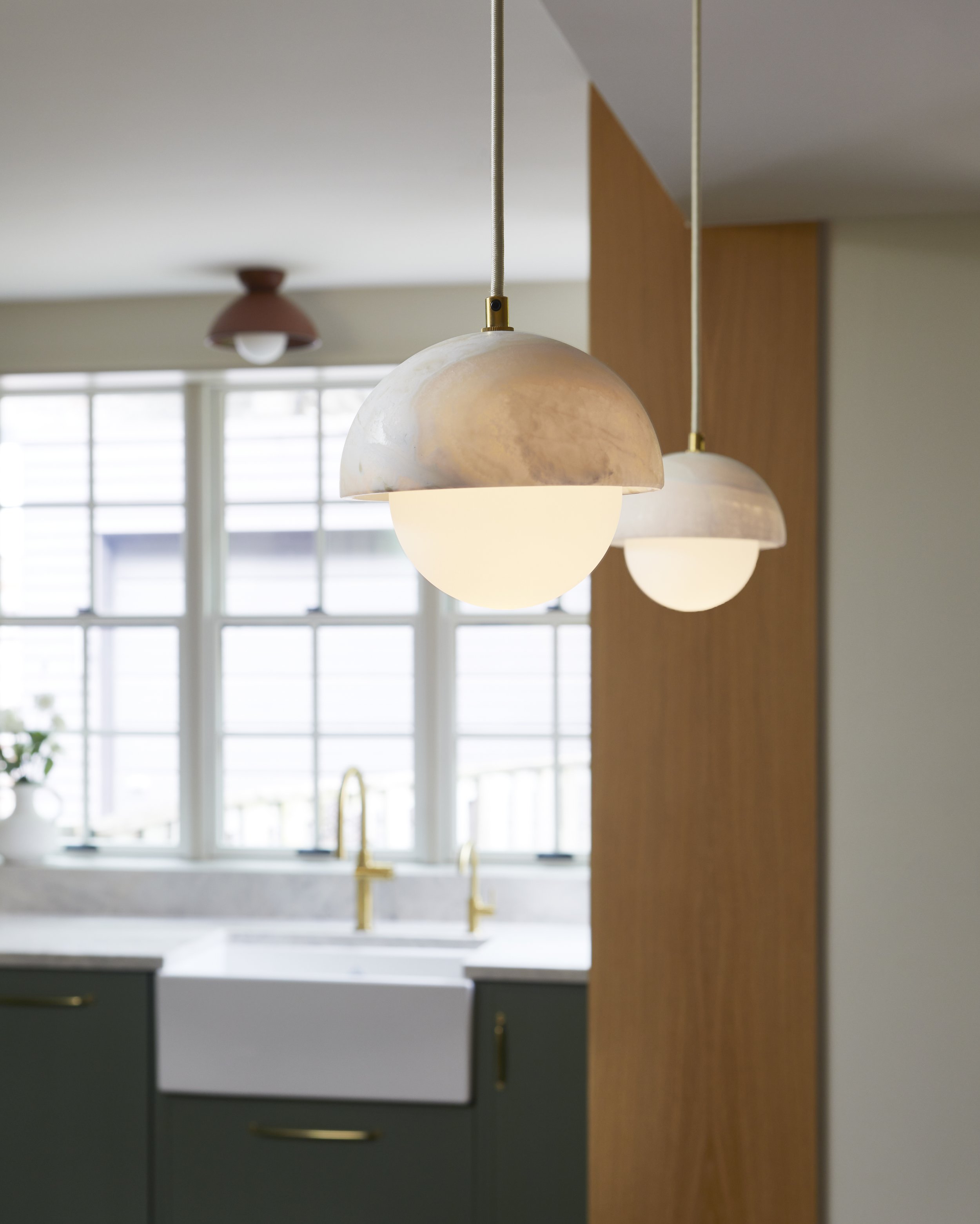 Close-up of two modern pendant lights with round white glass shades hanging in a kitchen with large windows, a white farmhouse sink, and dark green cabinets with gold hardware.