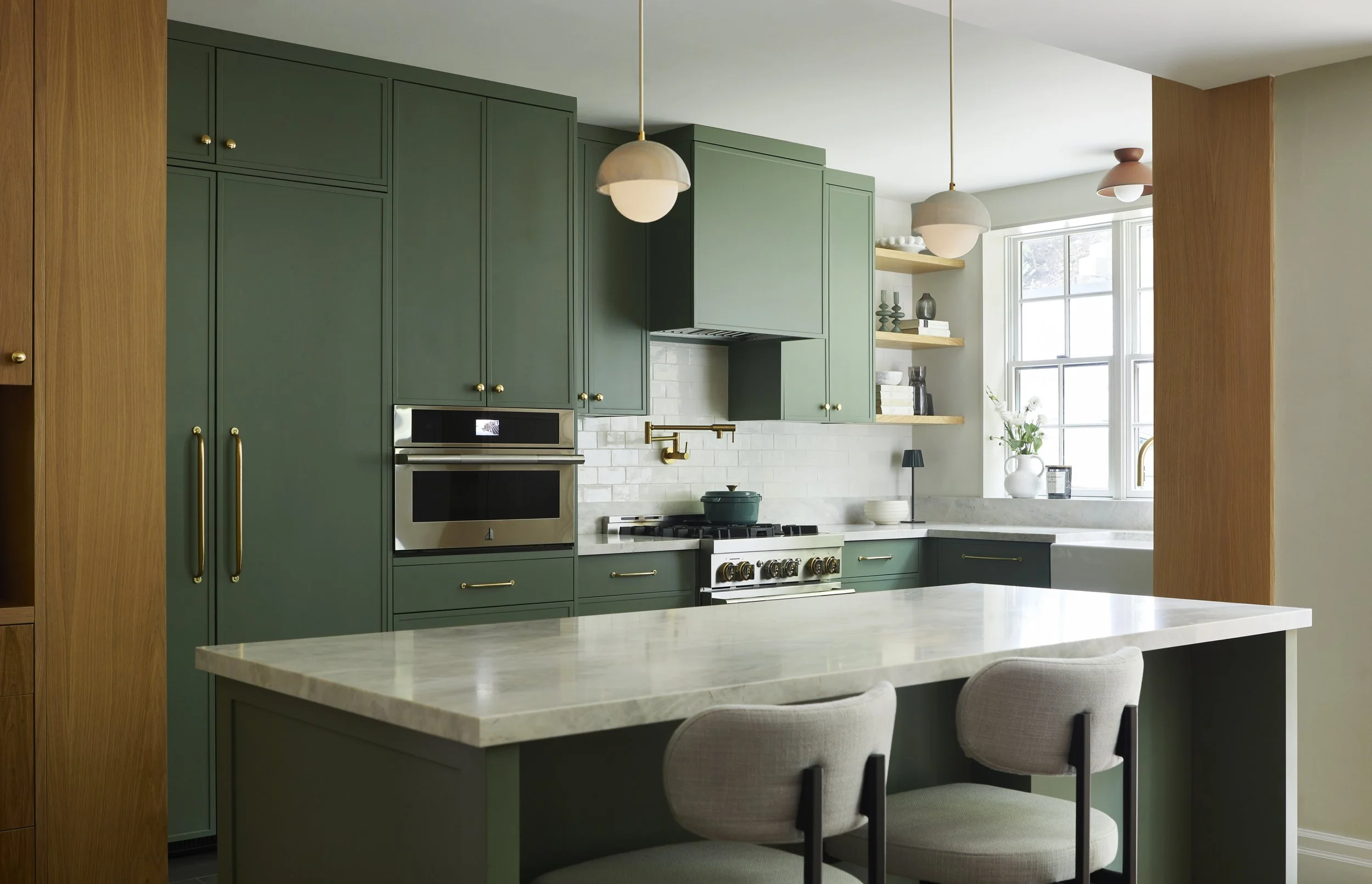 Modern kitchen with green cabinetry, a white marble island, and open wooden shelving near a window, decorated with vases and small decor items.