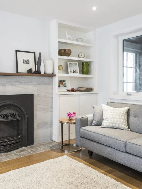 A cozy living room corner with a gray sofa, decorative pillows, a round wooden side table with pink flowers, built-in white shelves holding decorative items, a fireplace with a black hearth, a window allowing natural light, and a beige rug on wooden flooring.