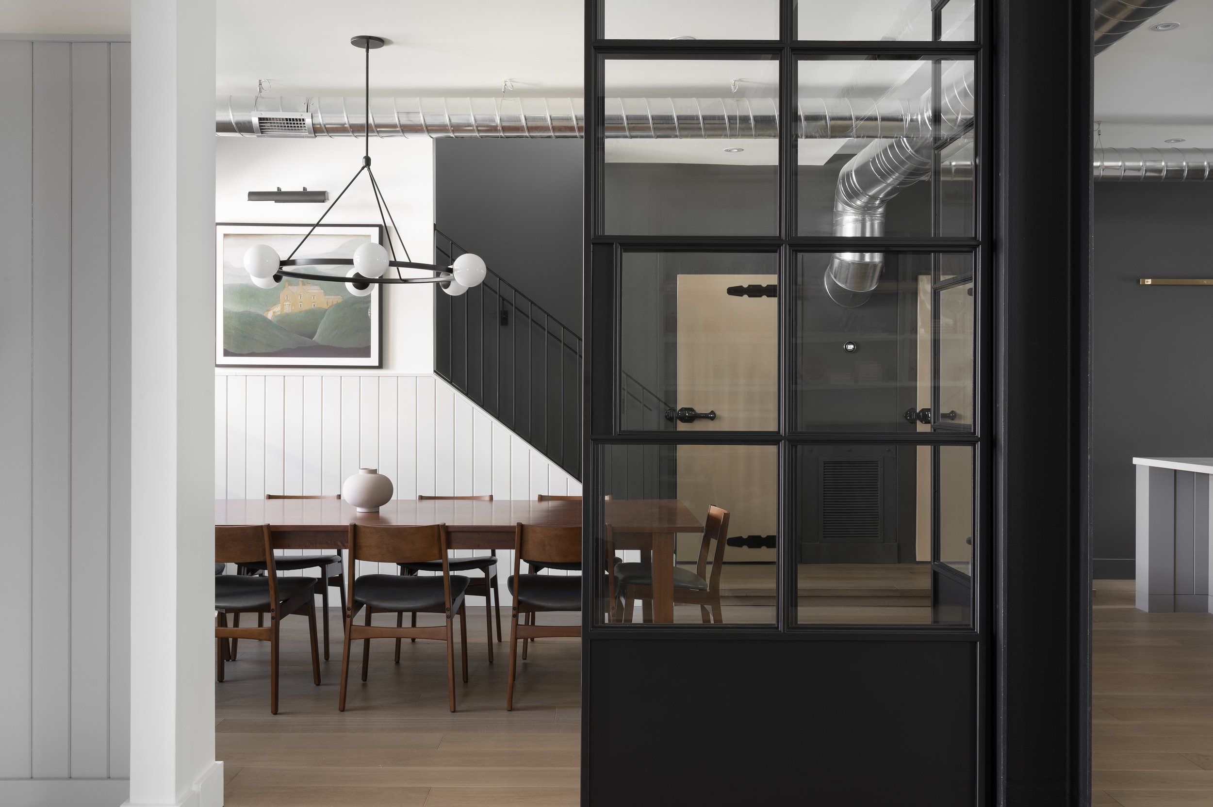 Modern dining room with wooden table, black chairs, white wall with paneling, and art painting, separated by a black framed glass partition, visible staircase, and industrial-style ductwork.
