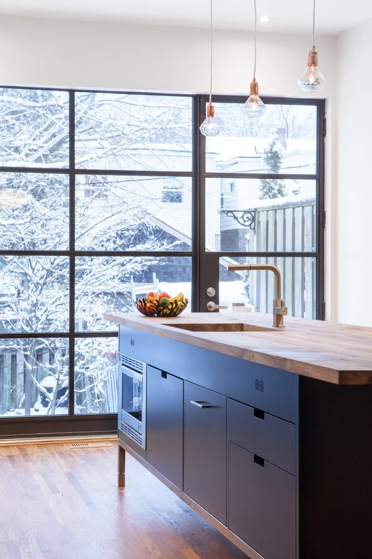 Modern kitchen with large window showing snowy outdoor scene, black cabinetry, and wooden countertop, hanging pendant lights.