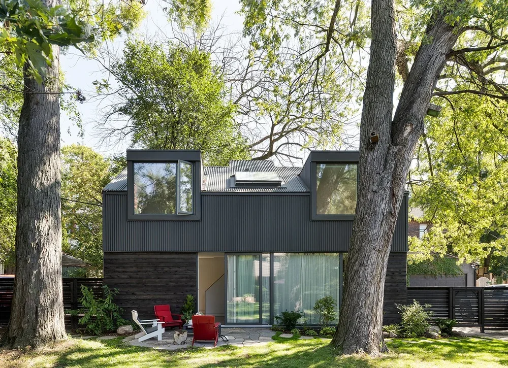 Modern two-story house with large windows, surrounded by tall trees and a fenced yard with a garden and outdoor seating area.
