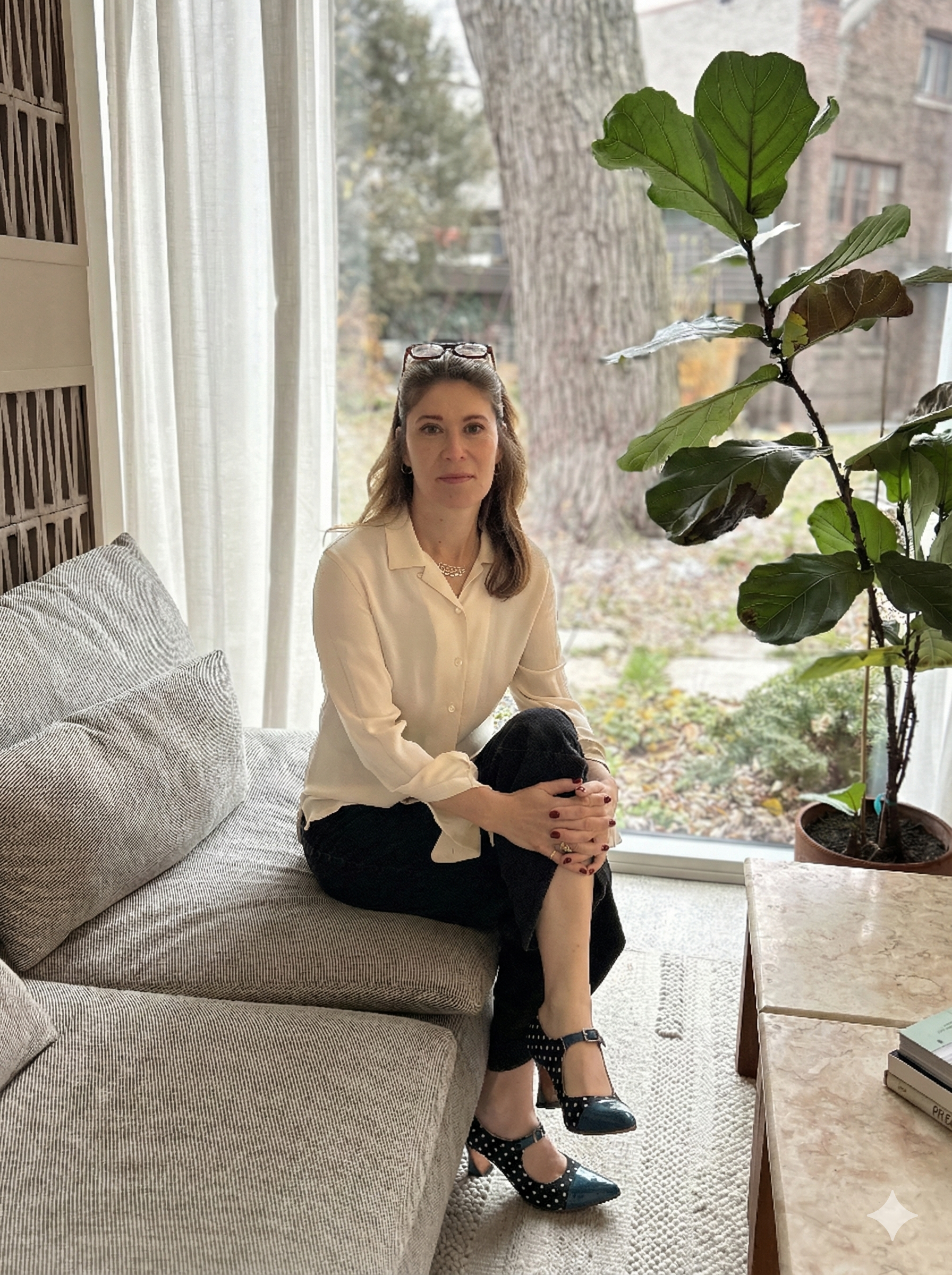 A woman with glasses on her head, wearing a cream blouse and black pants, sitting on a gray striped couch with her legs crossed, in a room with large windows and white curtains, next to a tall potted plant and a marble-topped table with books.