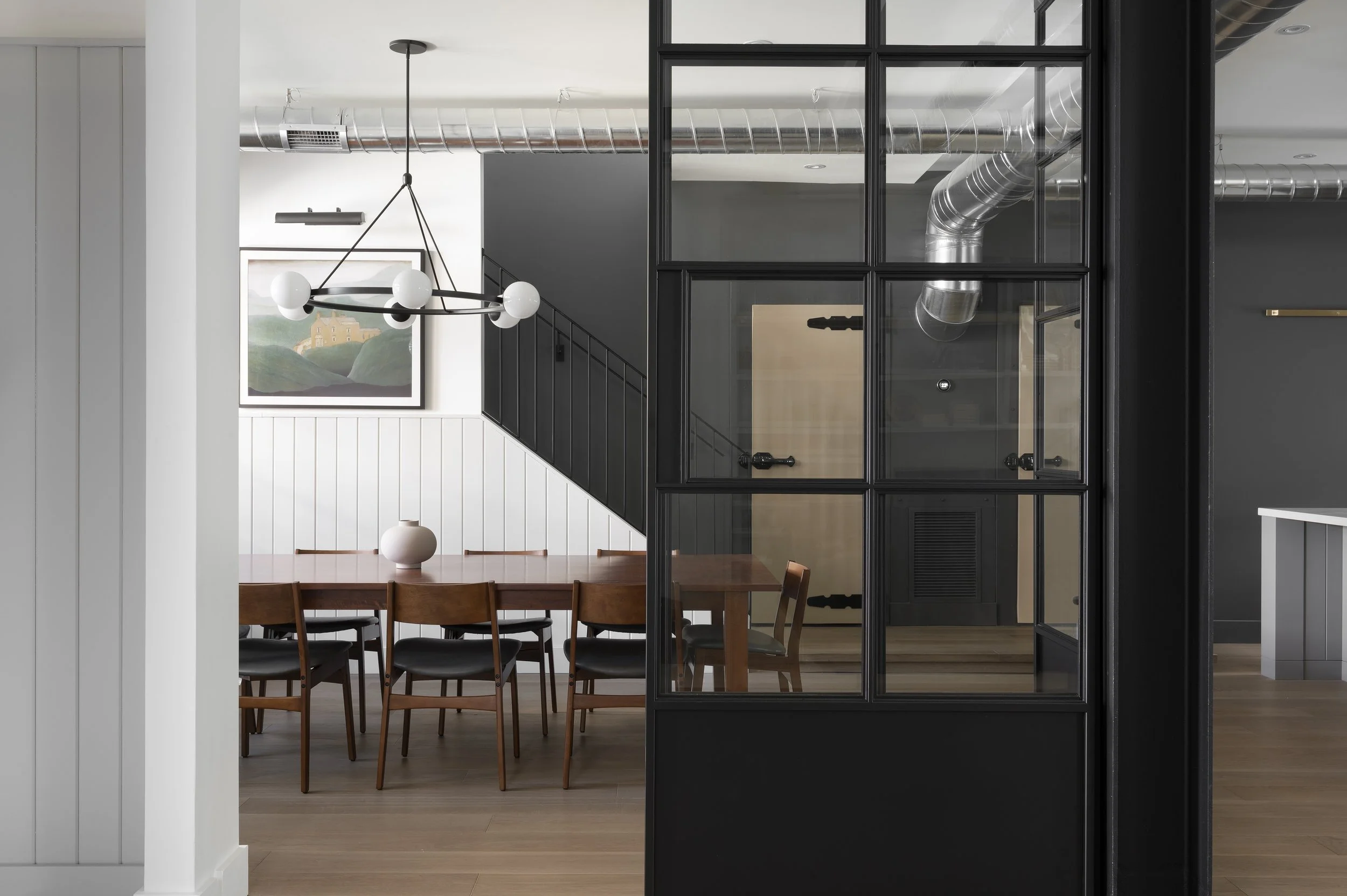 Interior of a modern dining area with a wooden table, six chairs, a white vase, and a black metal and glass wall divider, with artwork and a staircase in the background.
