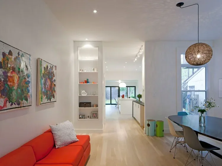 Open-plan modern living space with a red sofa, abstract artwork on the wall, white shelves with decor, a dining table with chairs, and a bright kitchen area with large windows.