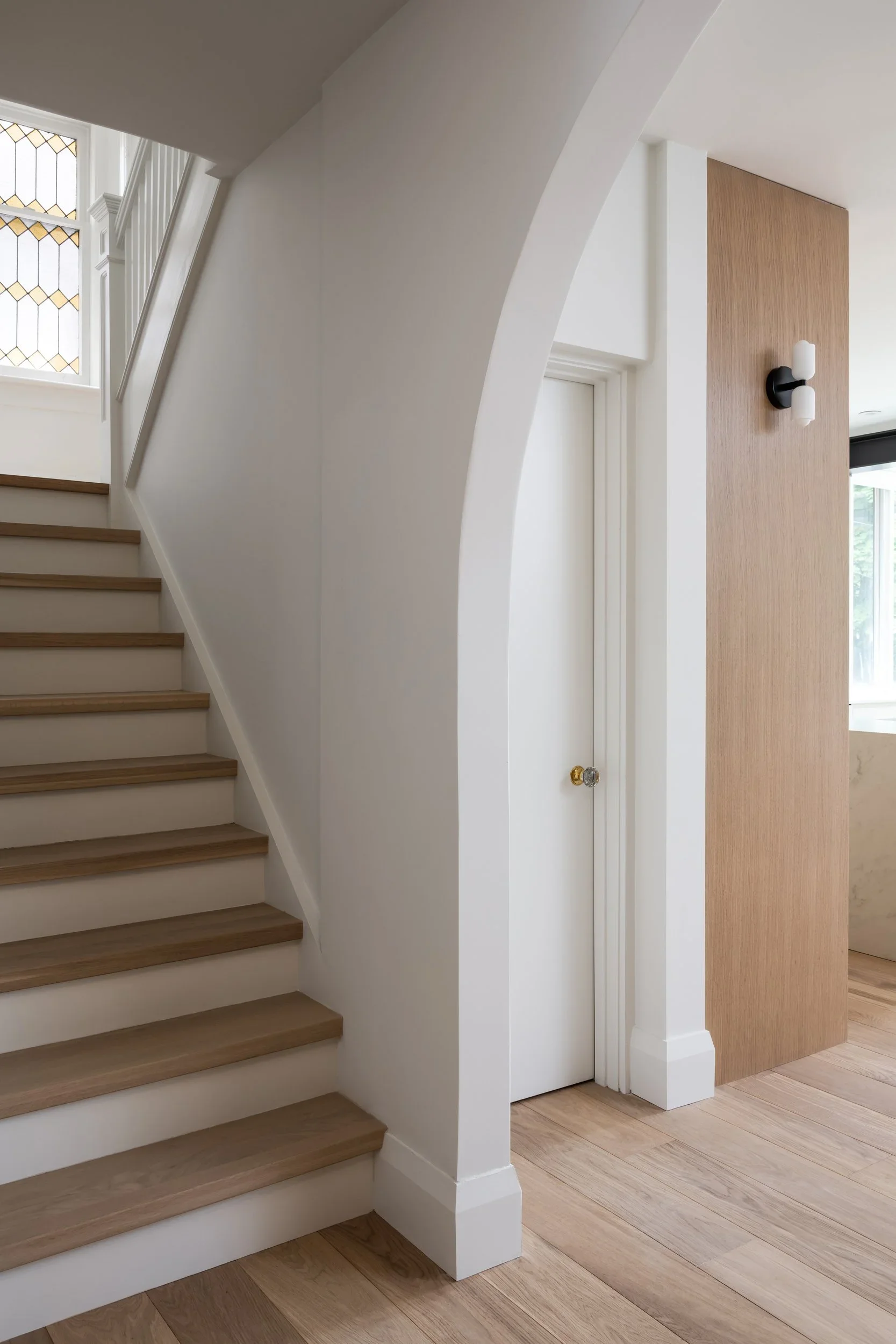 Interior view of a modern house showing wooden stairs, a white door with a brass knob, and a wooden wall panel with a decorative black and white double sconce light fixture.