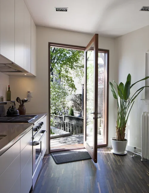 Open door leading to outdoor deck with trees and wooden fencing, inside a modern kitchen with white cabinets, a potted plant, and hardwood floors.