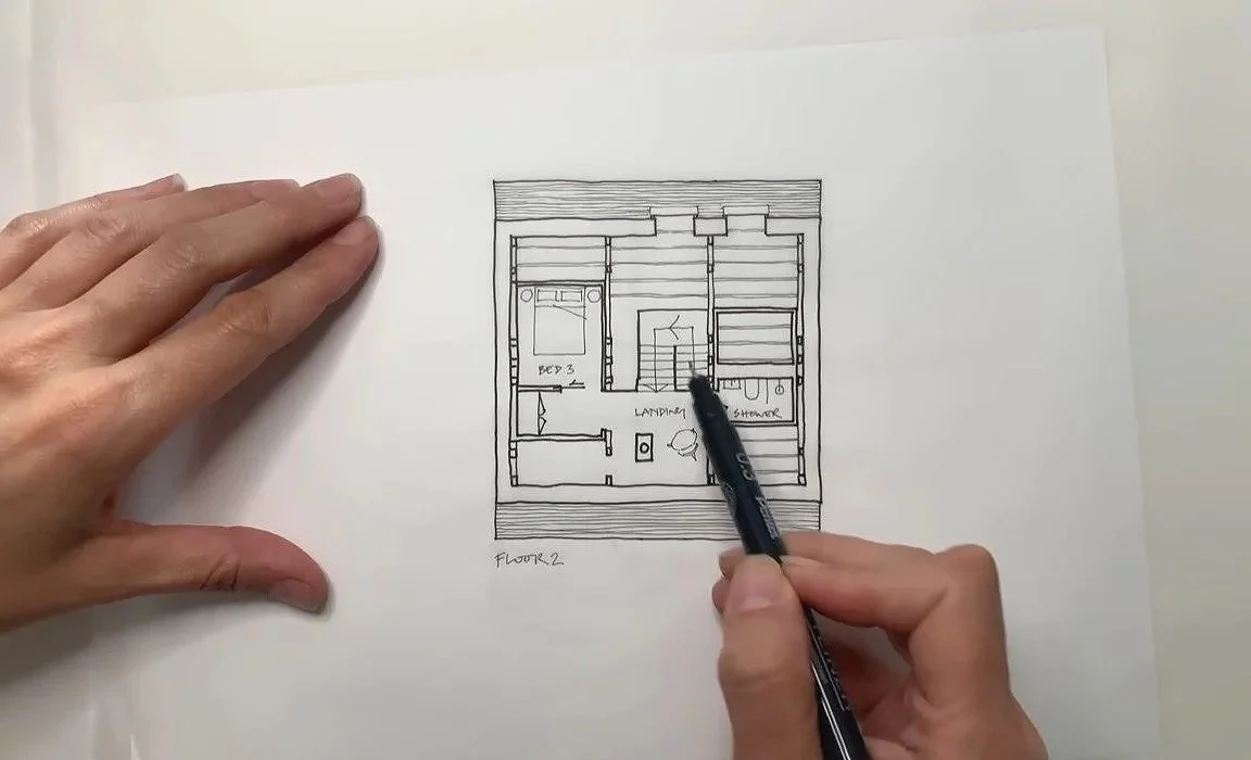 A person drawing a floor plan of a house on white paper with a black pen, showing a bedroom labeled 'Bed 3', a landing area, and a bathroom with shower.