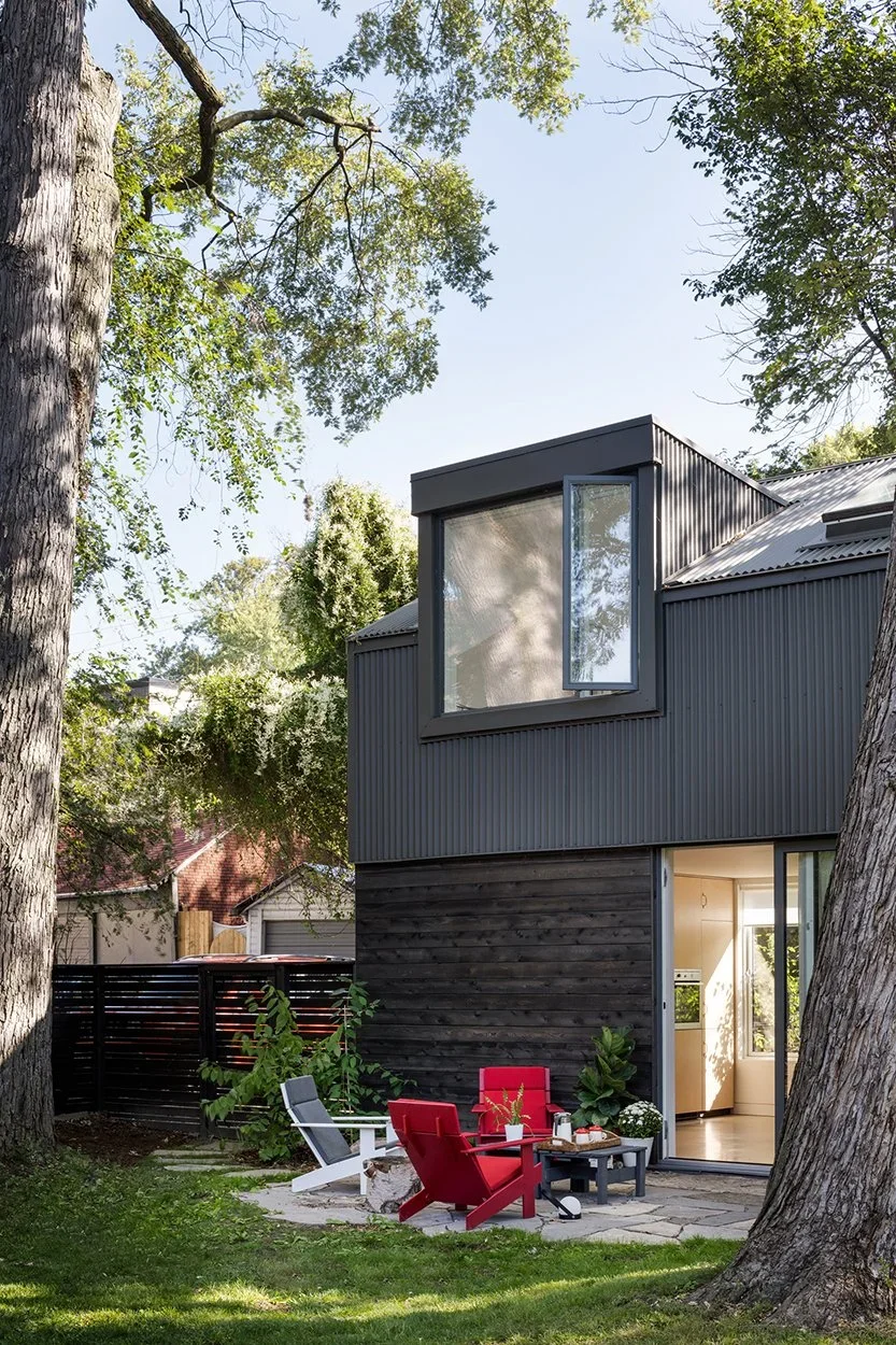 Modern two-story house with black and gray exterior, large open window on upper level, sliding glass door open to porch with red and gray outdoor chairs, surrounded by trees and greenery.