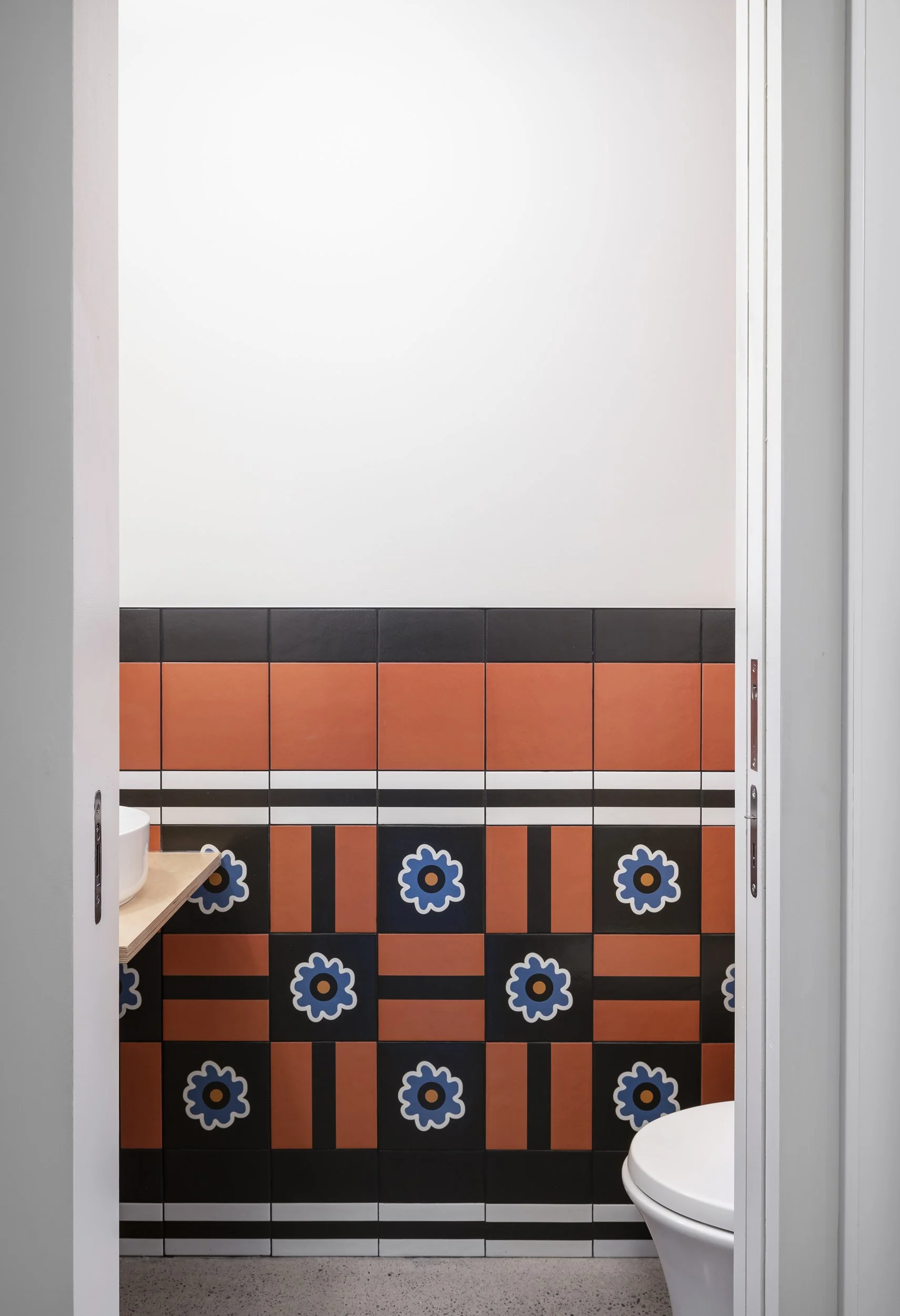 An inside view of a bathroom with colorful floral and geometric patterned tiles on the wall, white upper wall, and a toilet visible in the lower right corner.