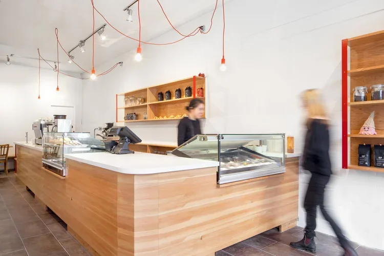 Inside a modern coffee shop with a wooden counter, a cash register, and desserts on display. Two blurred customers are at the counter, with a wooden shelf holding jars and bags behind them, and three pendant lights hanging from the ceiling.