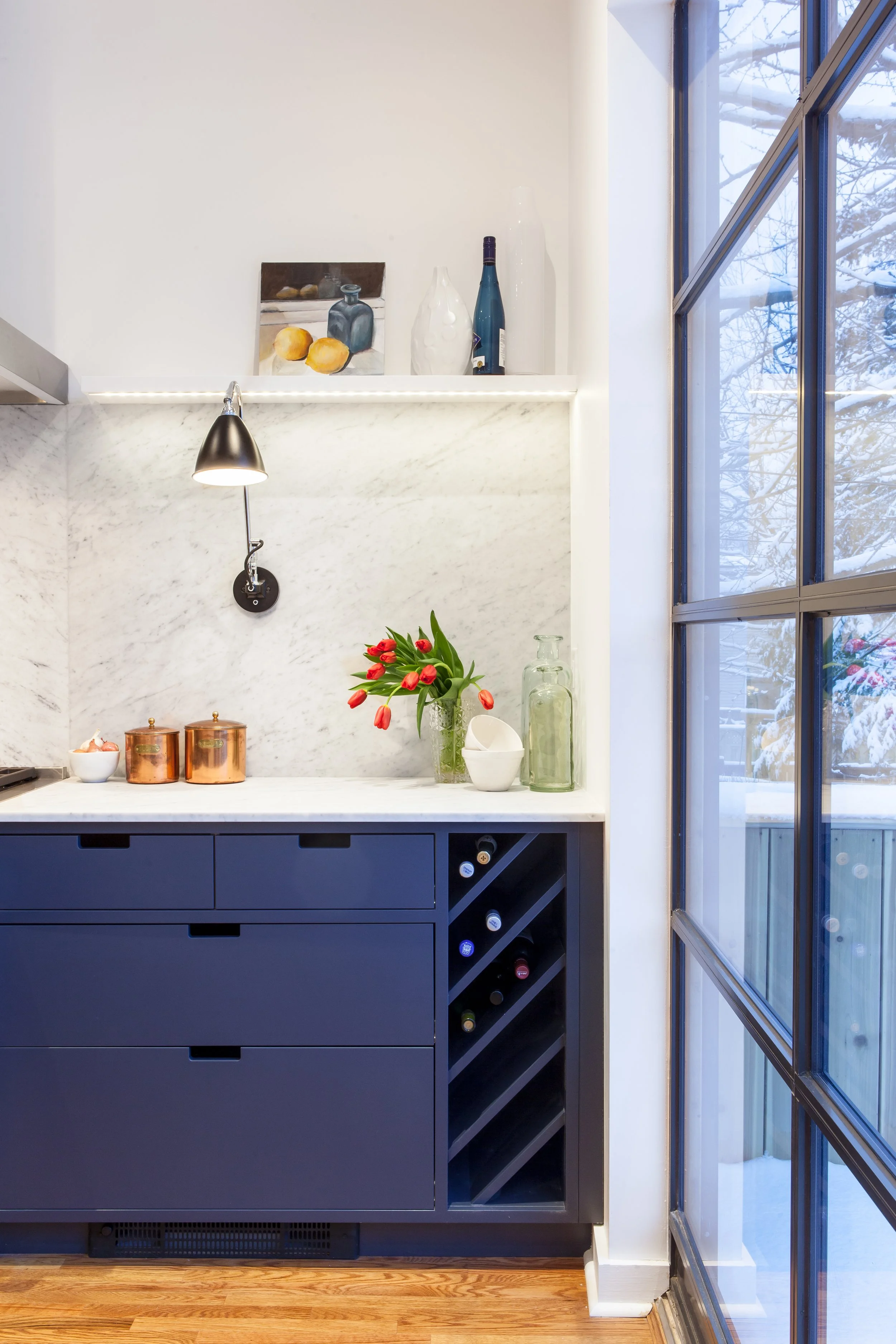 Interior view of a modern kitchen with a dark blue cabinet, a white marble countertop, and a marble backsplash. Decor items include a vase with red tulips, glass bottles, and small copper containers. A black wall-mounted lamp is above the countertop. Large windows show snow outside.