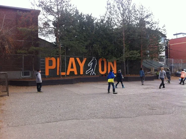 Children playing near a sign that says 'PLAY ON' with a figure of a person playing a sport, in an outdoor area with trees and buildings in the background.