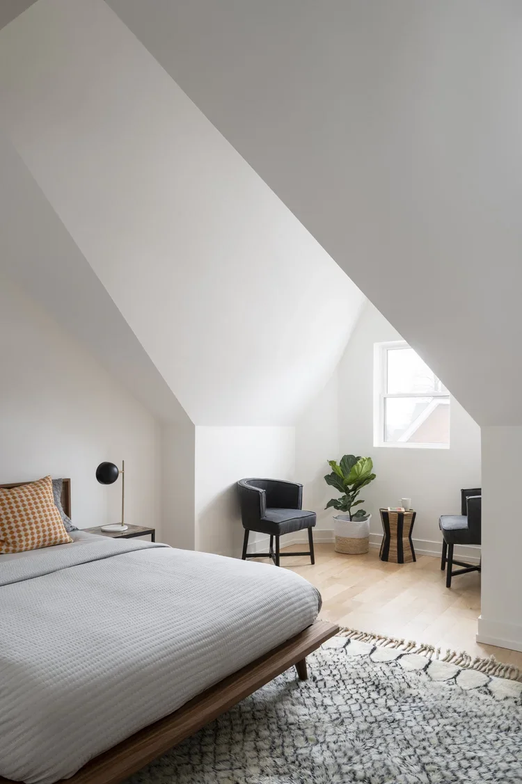 Bedroom with sloped ceiling, white walls, wooden floor, a bed with white bedding and an orange patterned pillow, a nightstand with a modern black lamp, two black chairs near a window, a large green potted plant, and a patterned rug.