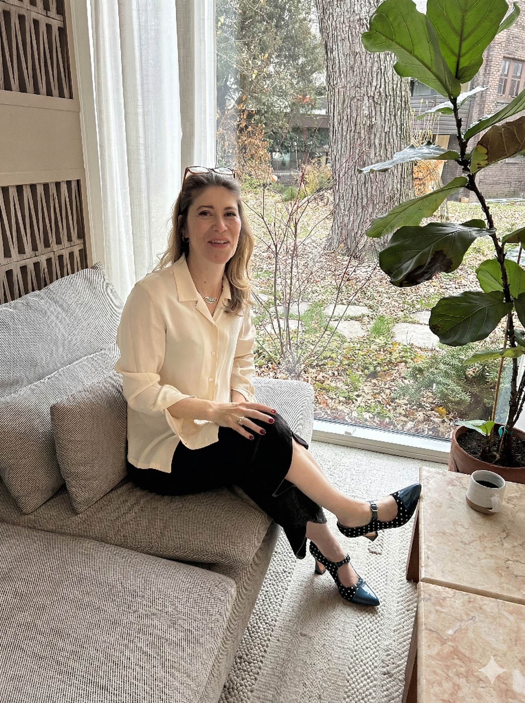 A woman sitting on a beige sofa in a bright room with large window, outdoor view of trees and buildings, holding her knee, wearing a cream blazer, black skirt, and polka-dot high heels, with a coffee cup on a table nearby.