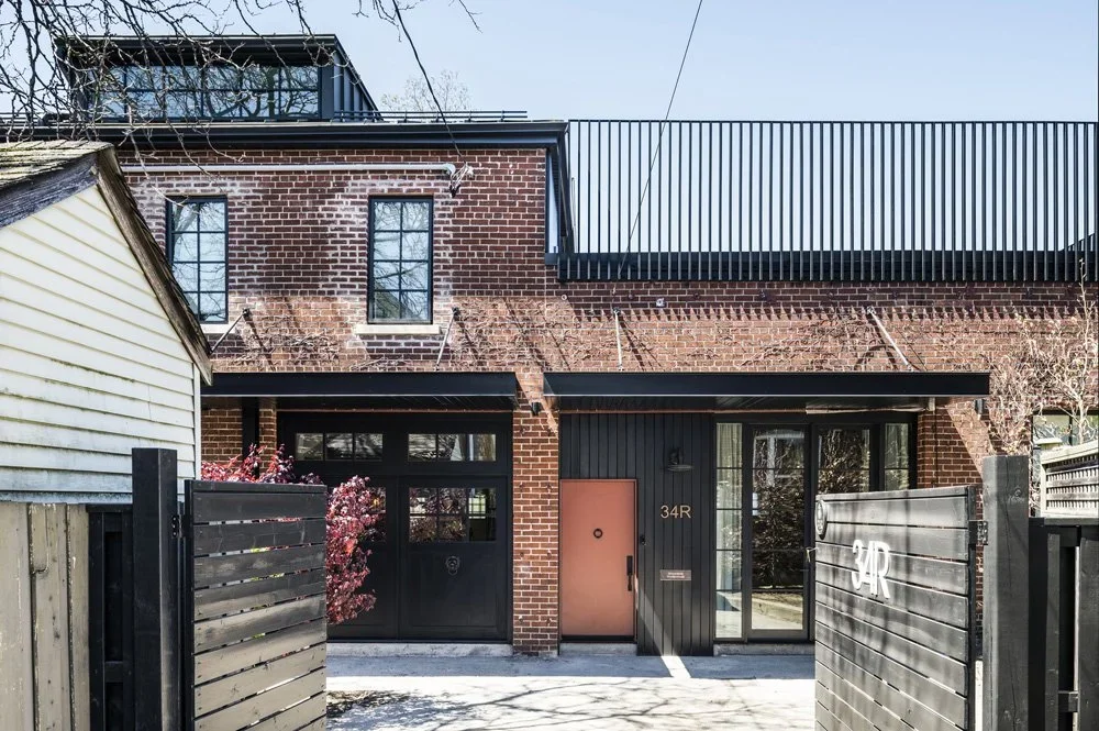 Modern townhouse with brick and dark paneling exterior, black gate with house number 34R, and a pink front door flanked by large windows. A small tree or shrub is in front of the house, and there is a neighboring house with a light-colored siding.
