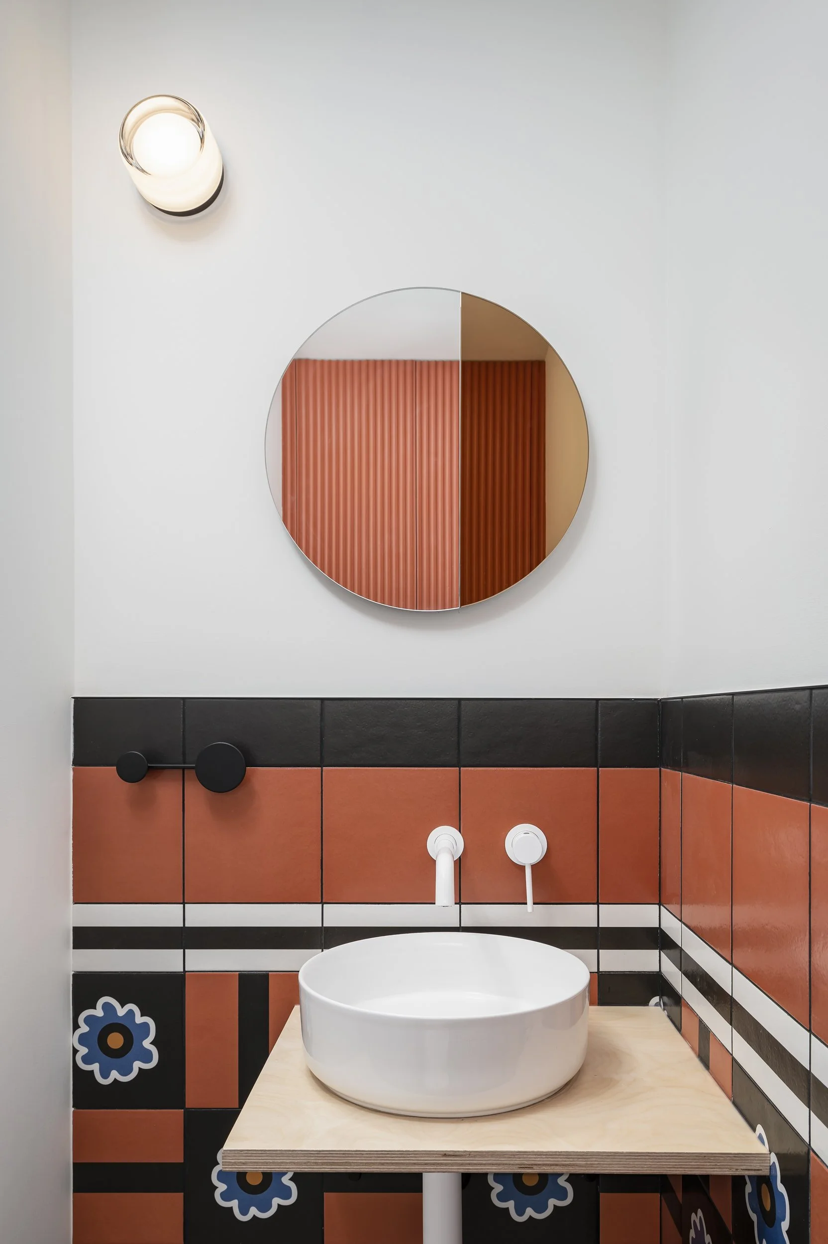 A modern bathroom with colorful tile walls, containing a round white vessel sink on a light wooden countertop, a circular mirror above, and a wall-mounted light fixture.