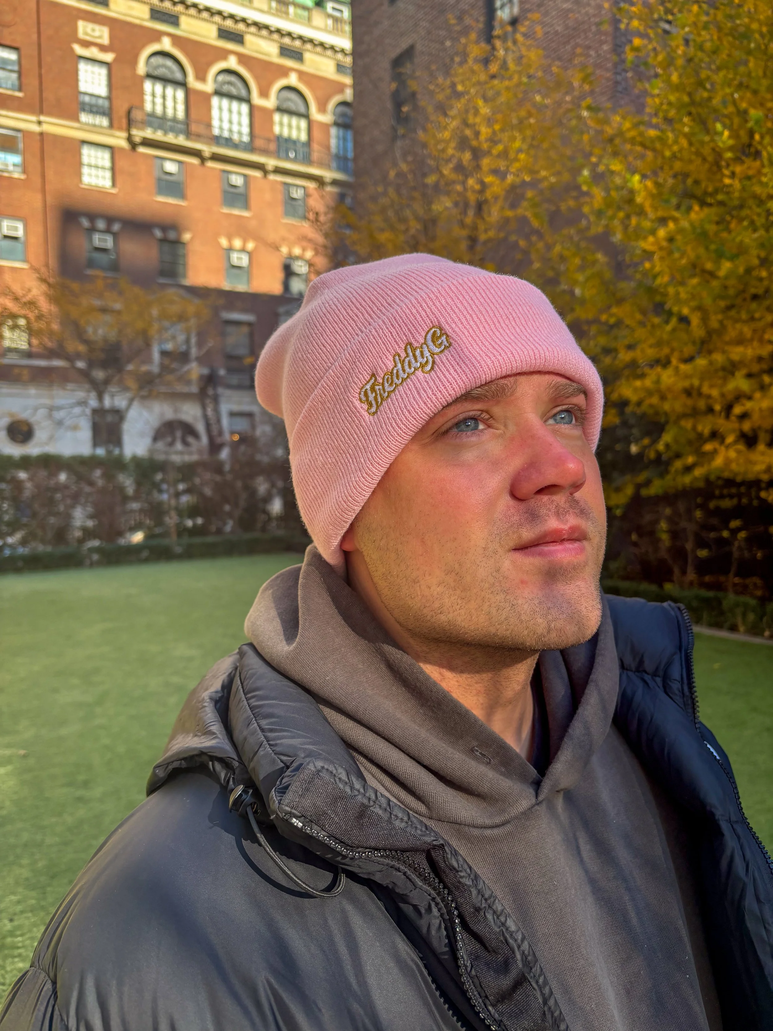 A man wearing a pink beanie with the embroidered words 'Freddy G' stands outdoors in front of autumn trees and brick buildings, looking contemplative.