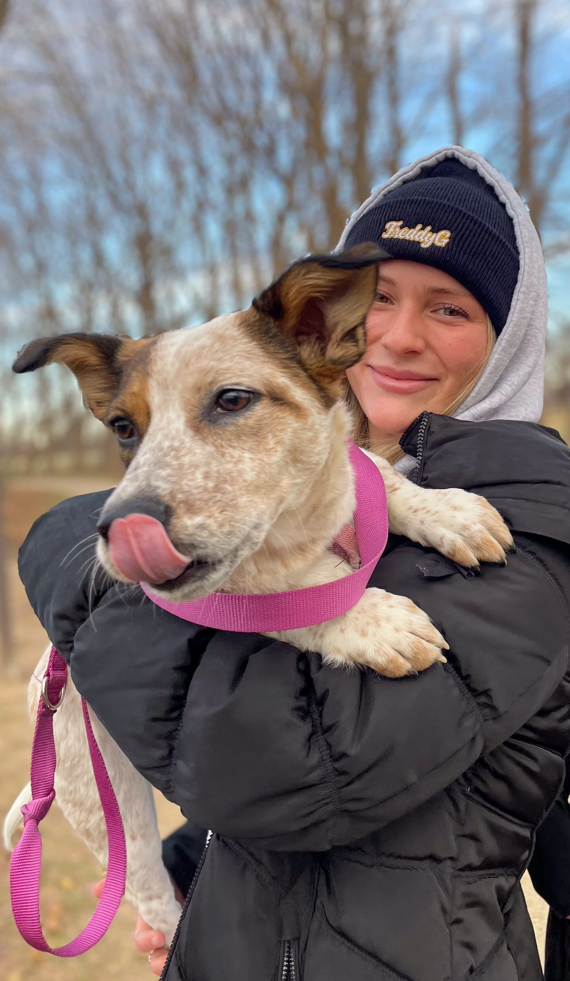 A woman in a hoodie that says 'Freddy G' and black jacket holding a small dog with a pink collar, outdoors with trees in the background.