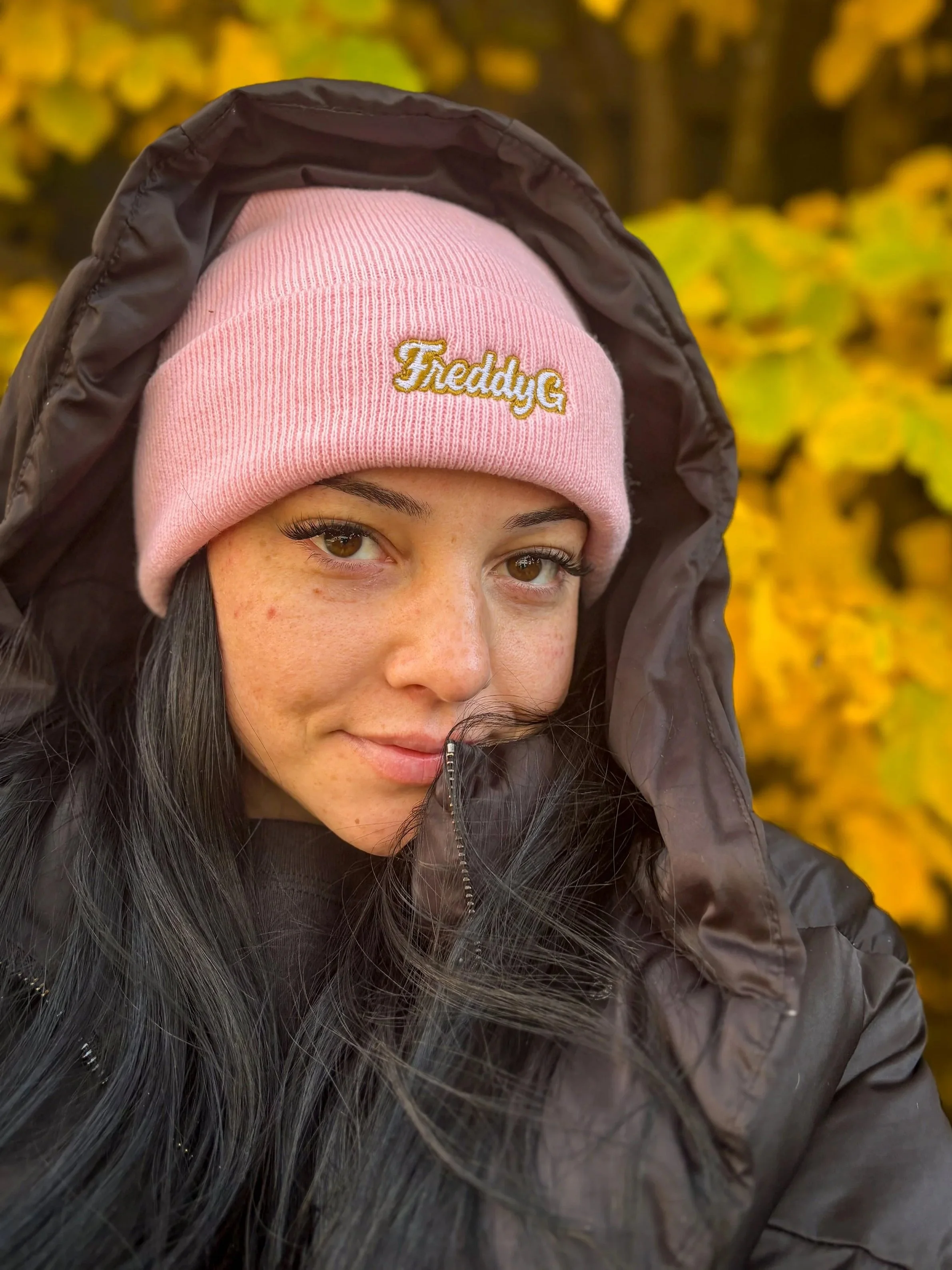 A woman outdoors wearing a pink beanie that says 'Freddy G' and a black jacket, with yellow autumn leaves in the background.