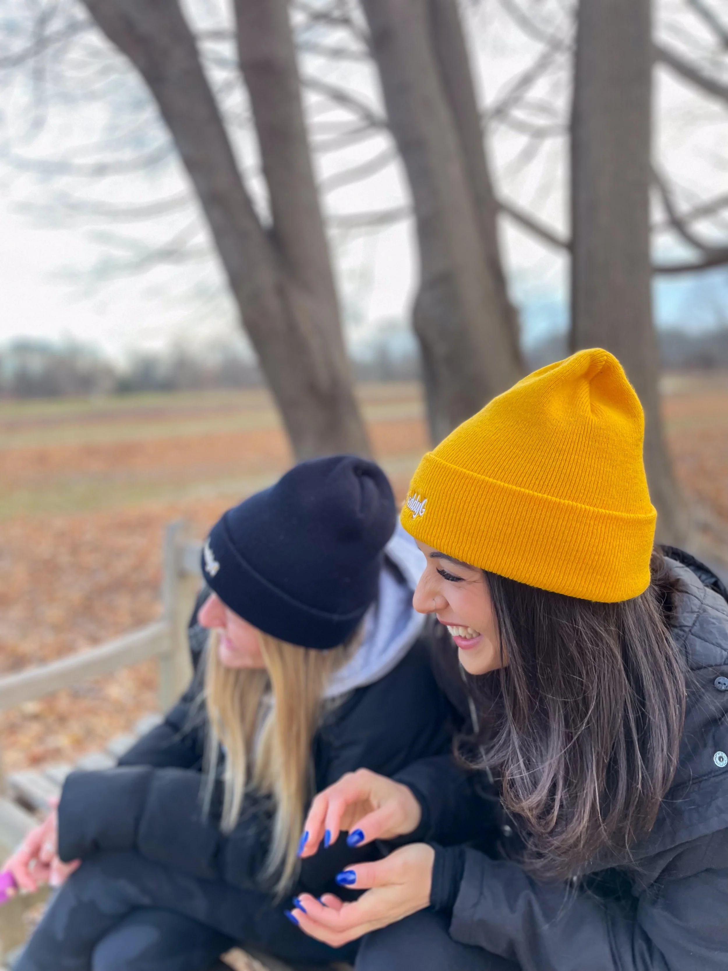 Two women sitting on a bench outdoors, wearing black jackets and 'Freddy G'  beanies, laughing and enjoying a moment together in a park with leafless trees in the background.