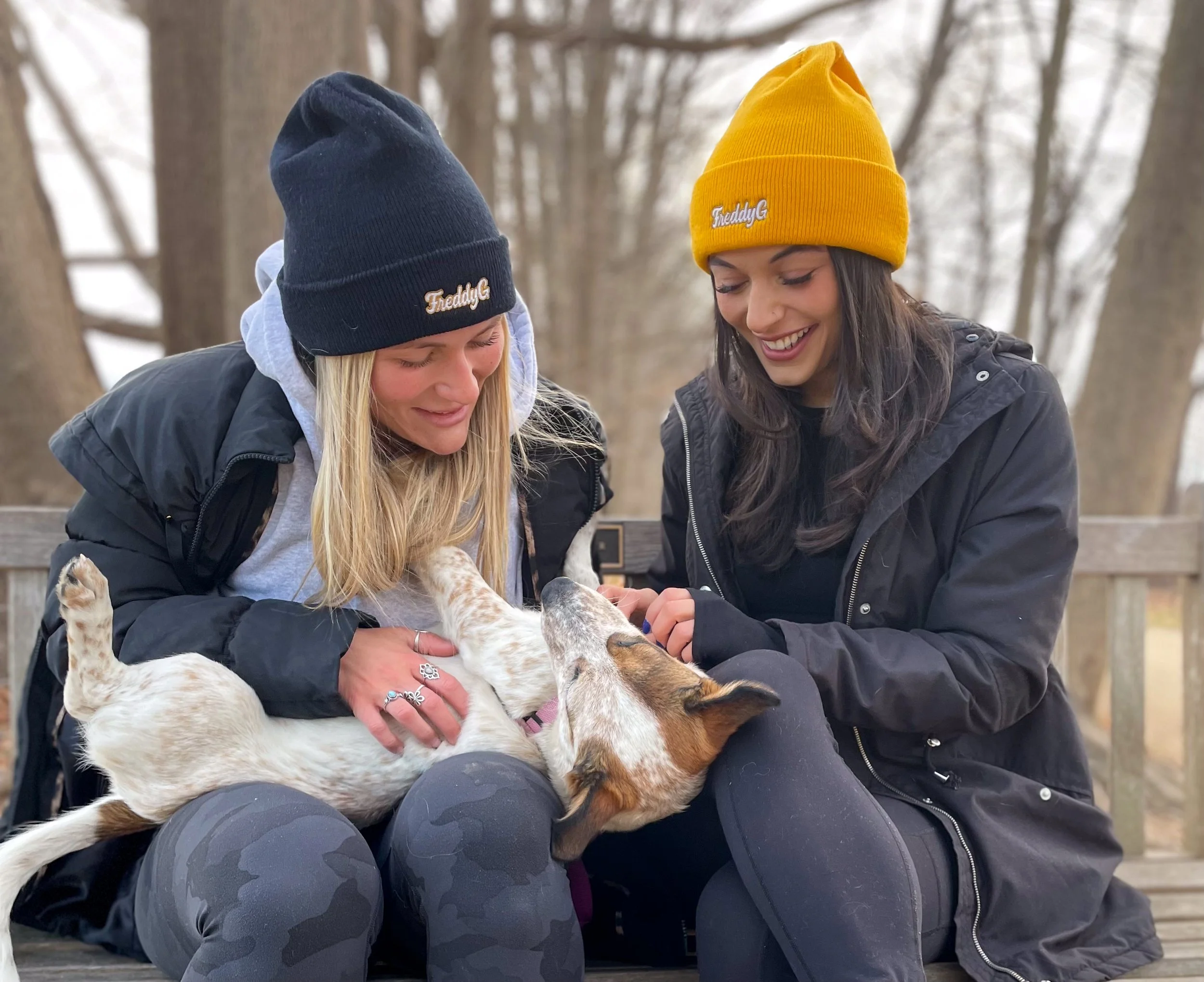 Two women wearing 'Freddy G' beanies and jackets sitting on a bench and playing with a small dog in a wooded outdoor area.