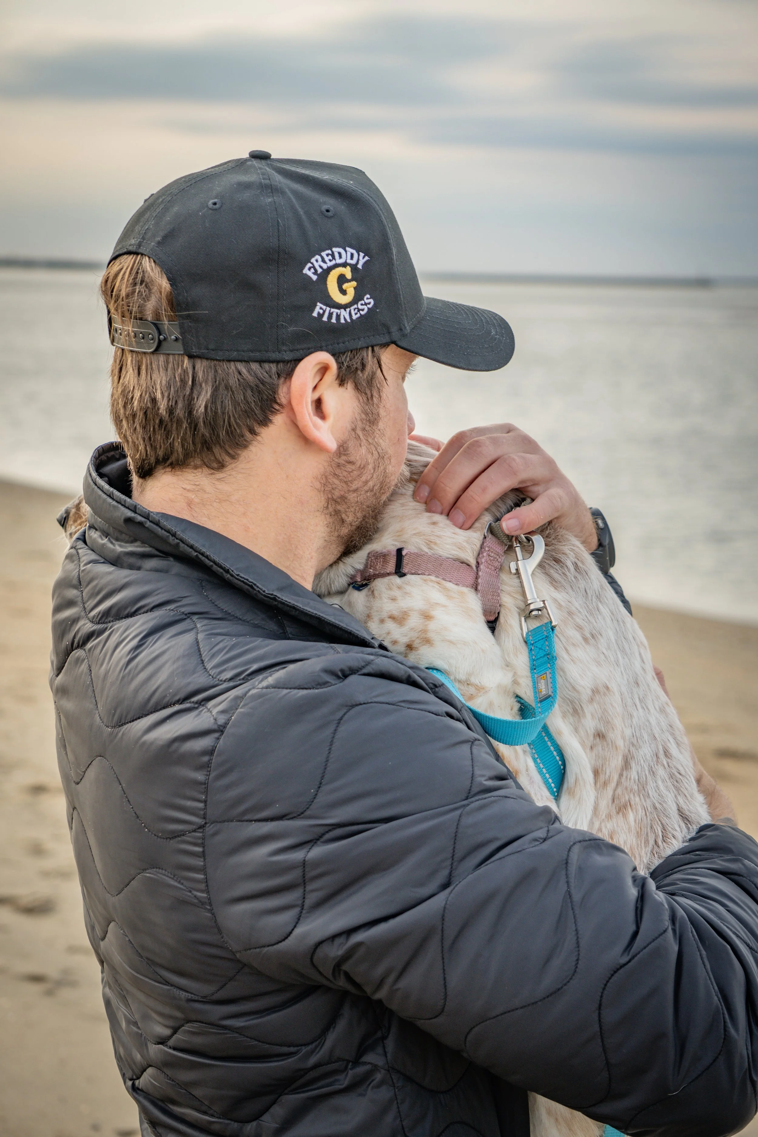 A man in a black 'Freddy G Fitness' cap and jacket hugging a white and brown dog on a beach near the water during cloudy weather.