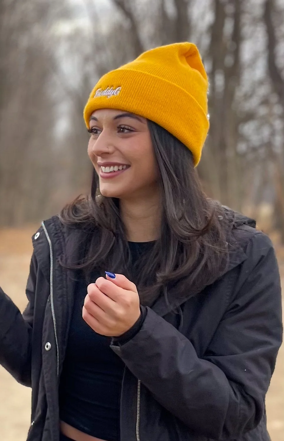 A young woman outdoors wearing a yellow 'Freddy G'  beanie and black jacket, smiling and looking to the side, with a blurred natural background.