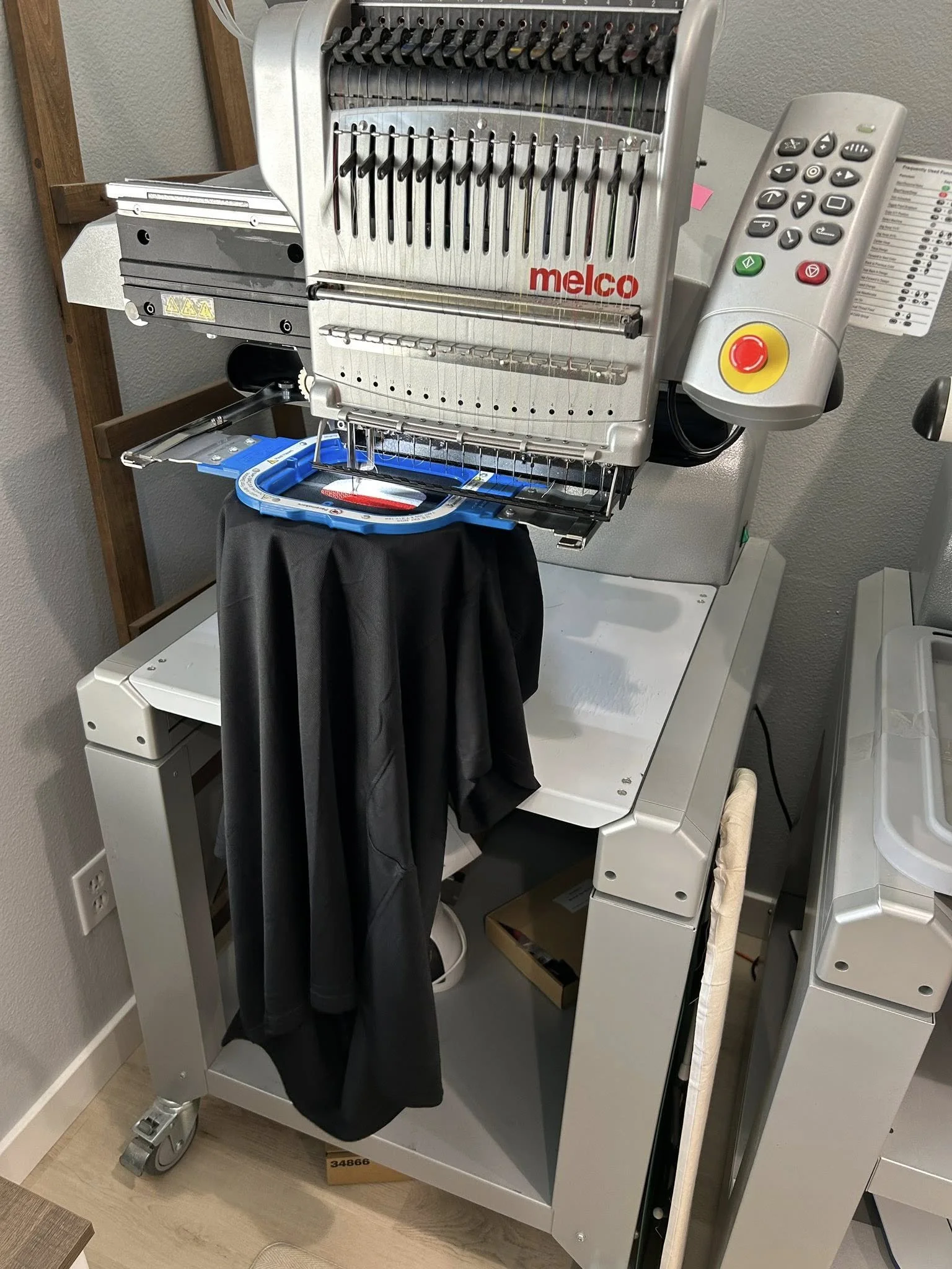 A commercial embroidery machine in a workspace, with black fabric draped over it, and a remote control attached to the machine.