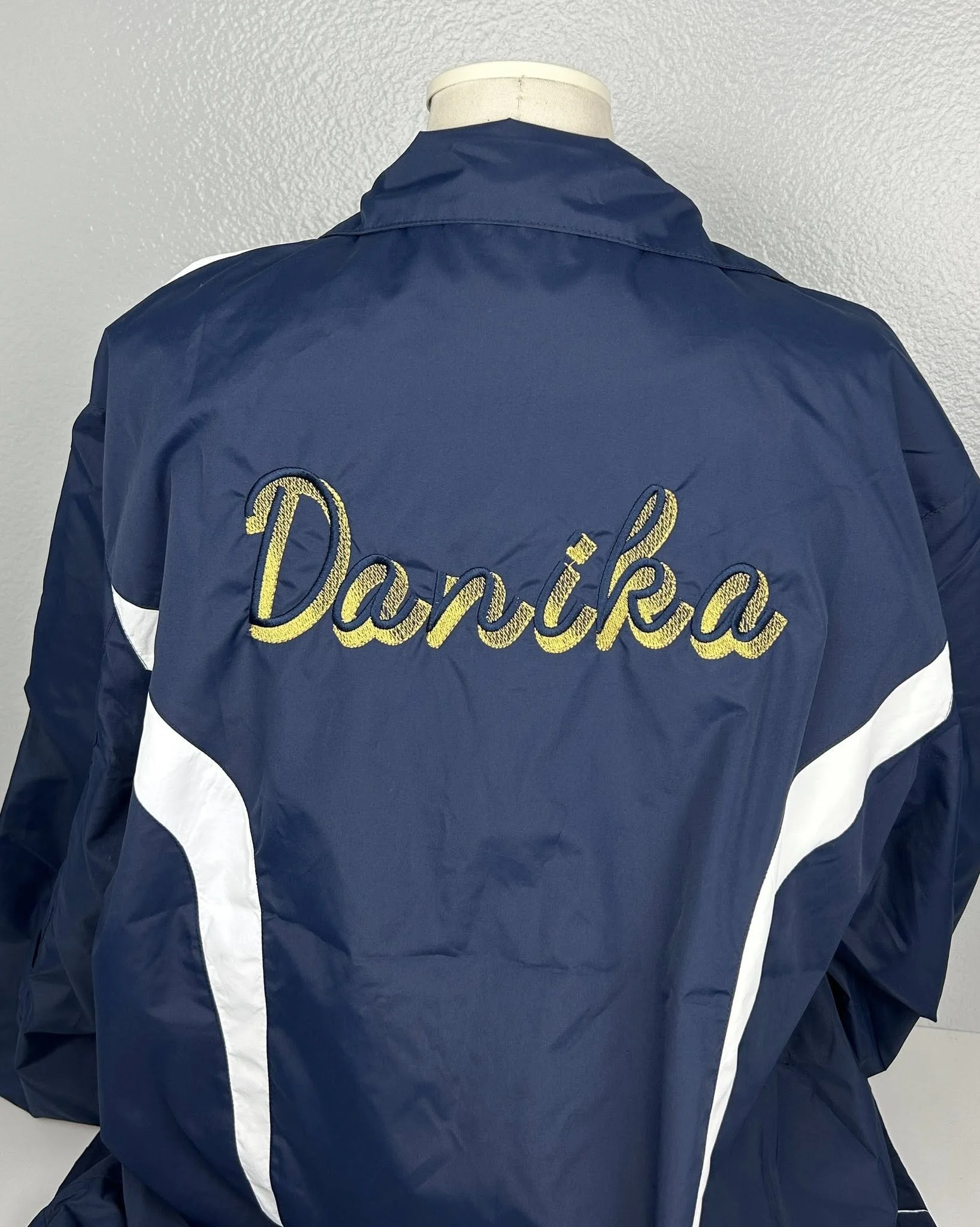 Back of a navy blue jacket with white stripes on the sleeves, embroidered with the name 'Danika' in yellow cursive on the back.