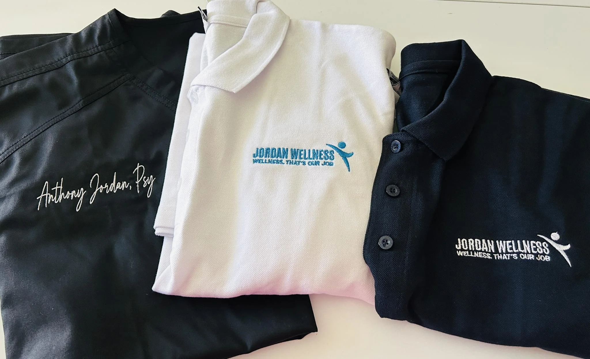 Three clothing items with embroidered logos: a black jacket with "Anthony Jordan, Psy" embroidered, a white polo shirt with "Jordan Wellness" logo, and a dark polo shirt with "Jordan Wellness Wellness, That's Our Job" embroidered.