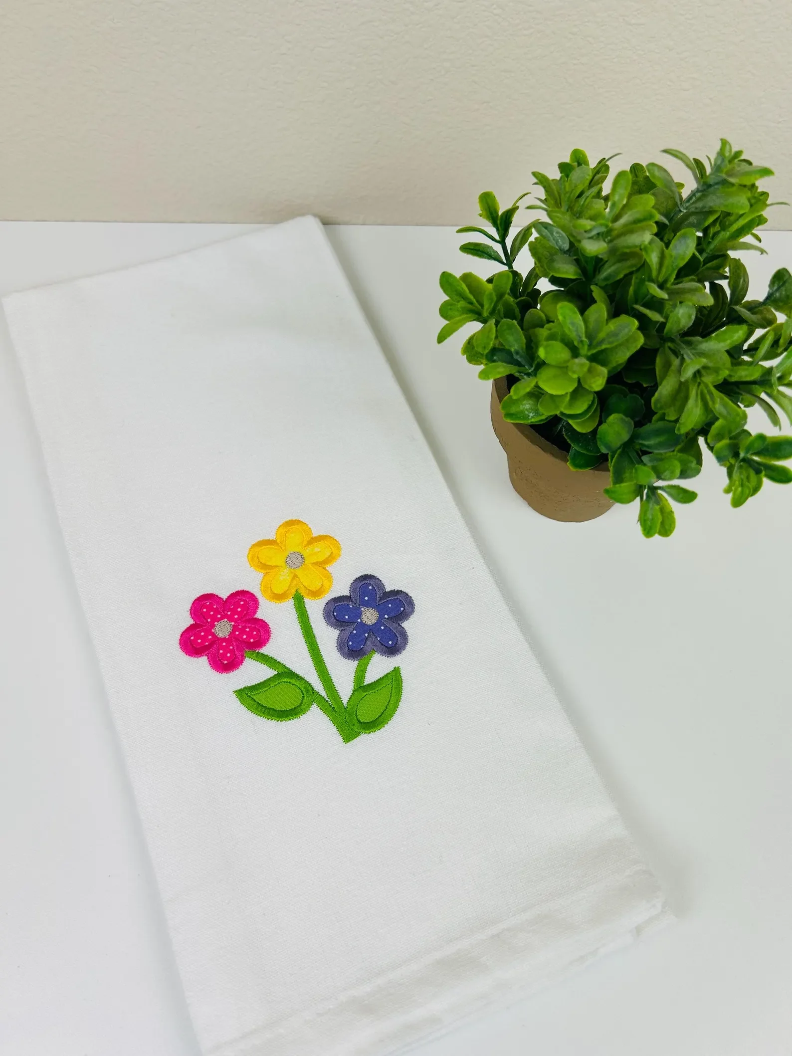 A white cloth napkin with colorful embroidered flowers in pink, yellow, and blue, and green leaves and stems, placed on a white surface next to a potted green plant with small, rounded leaves.