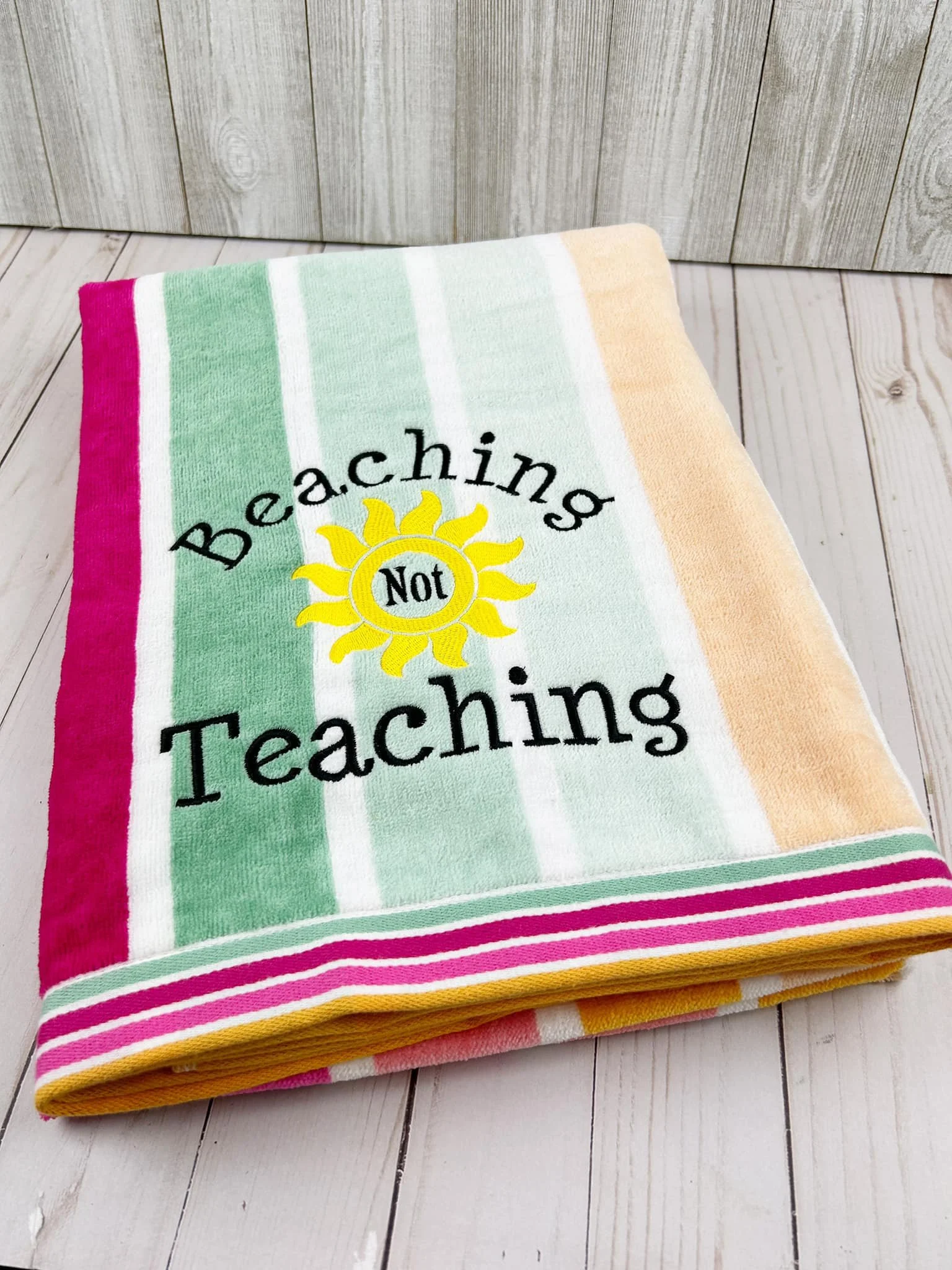 Colorful beach towel with the words 'Beaching Not Teaching' embroidered on it, featuring a yellow sun with the word 'Not' inside.