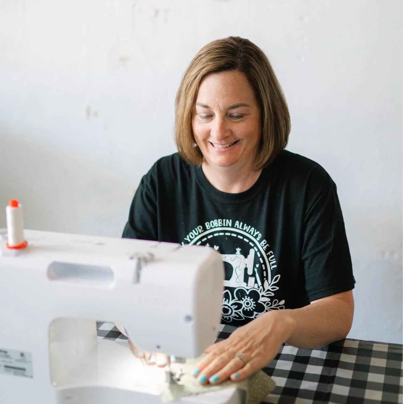 A woman with shoulder-length brown hair sewing on a white sewing machine, smiling while working. She wears a black T-shirt with a white graphic and text. The background is plain and light-colored.