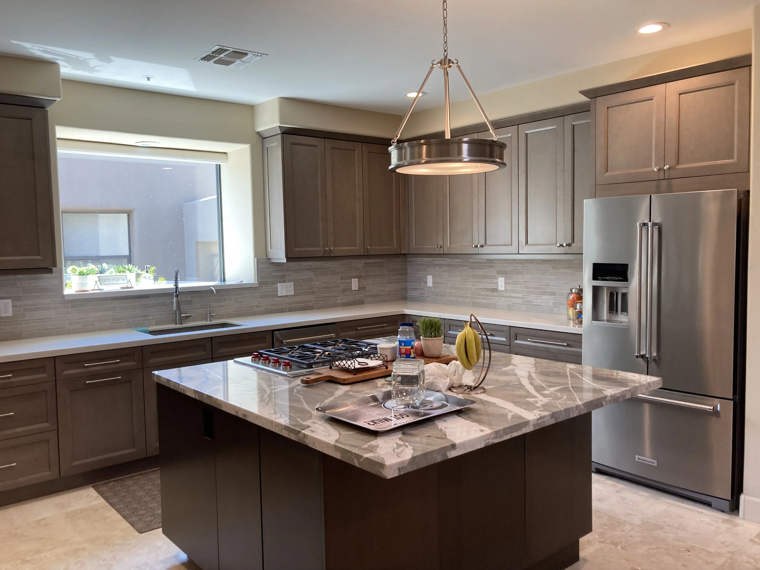 Modern kitchen with gray cabinets, marble island, stainless steel refrigerator, and a large window above the sink.