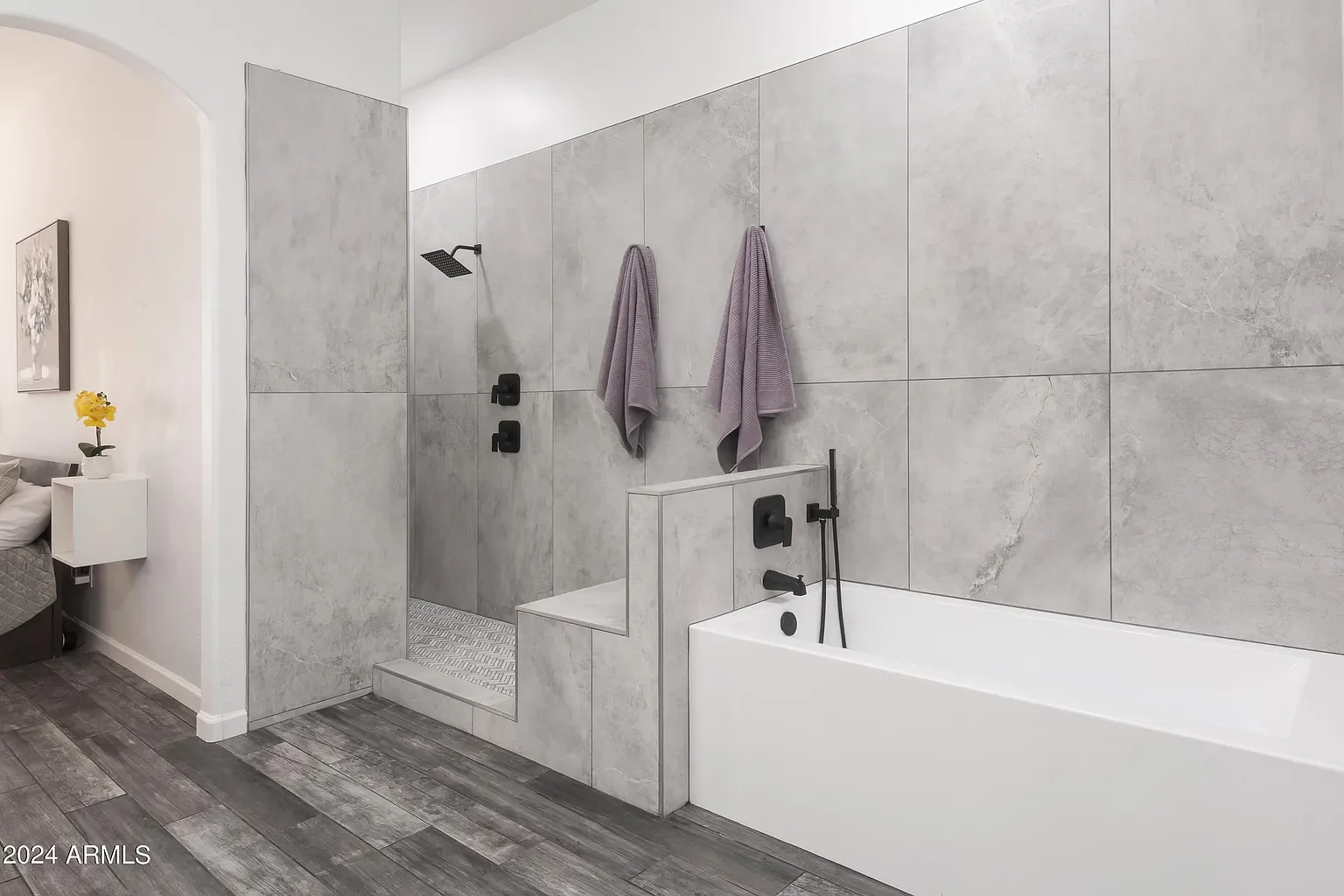 Modern bathroom with gray tiled wall, bathtub, and walk-in shower area separated by a low wall. The shower has black fixtures and two hanging purple towels.