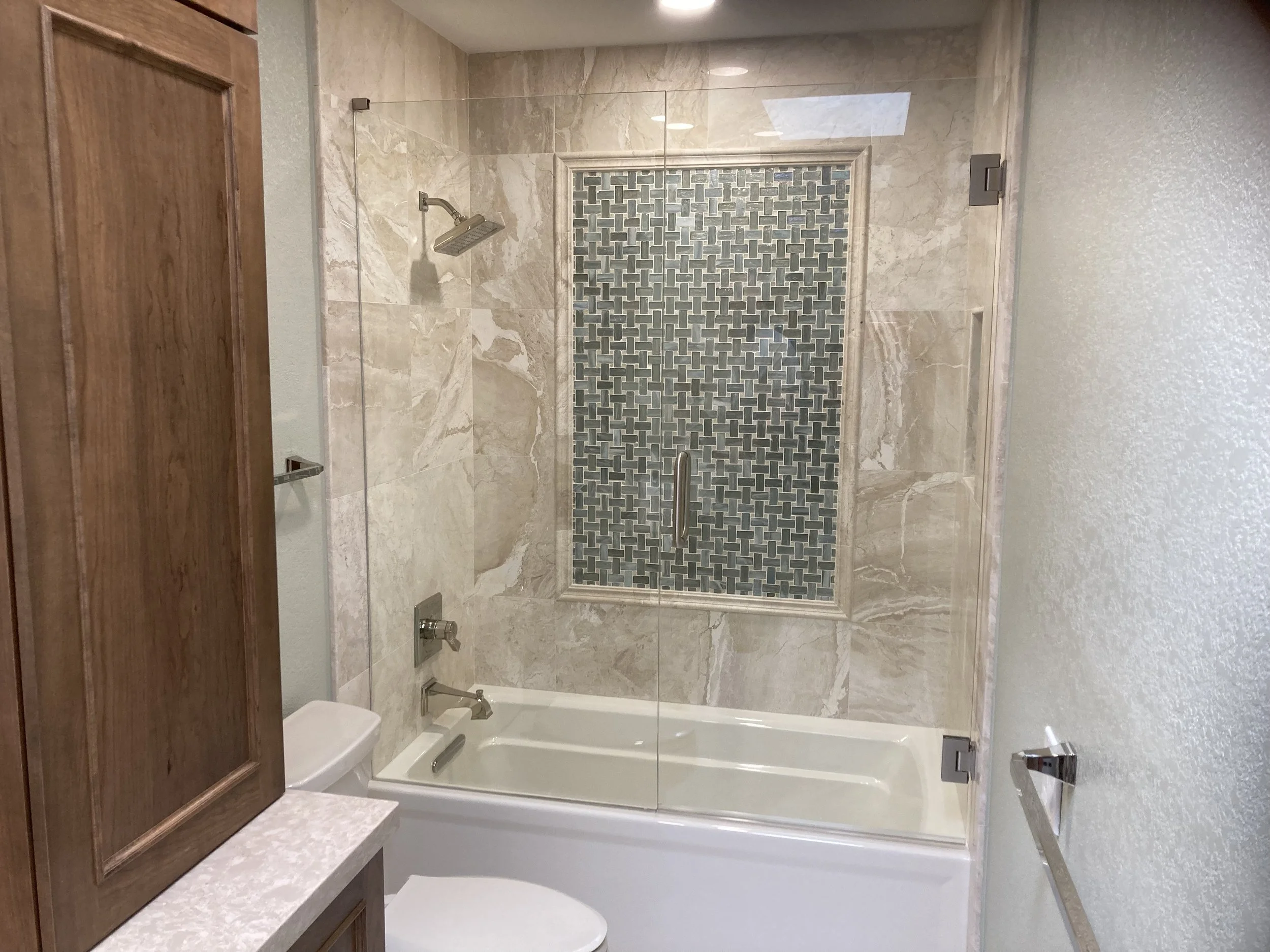 Bathroom with a bathtub and shower, glass shower door, beige marble tiles, a niche with patterned blue and gray tiles, vanity with wooden cabinet, white countertop, toilet, towel bar.