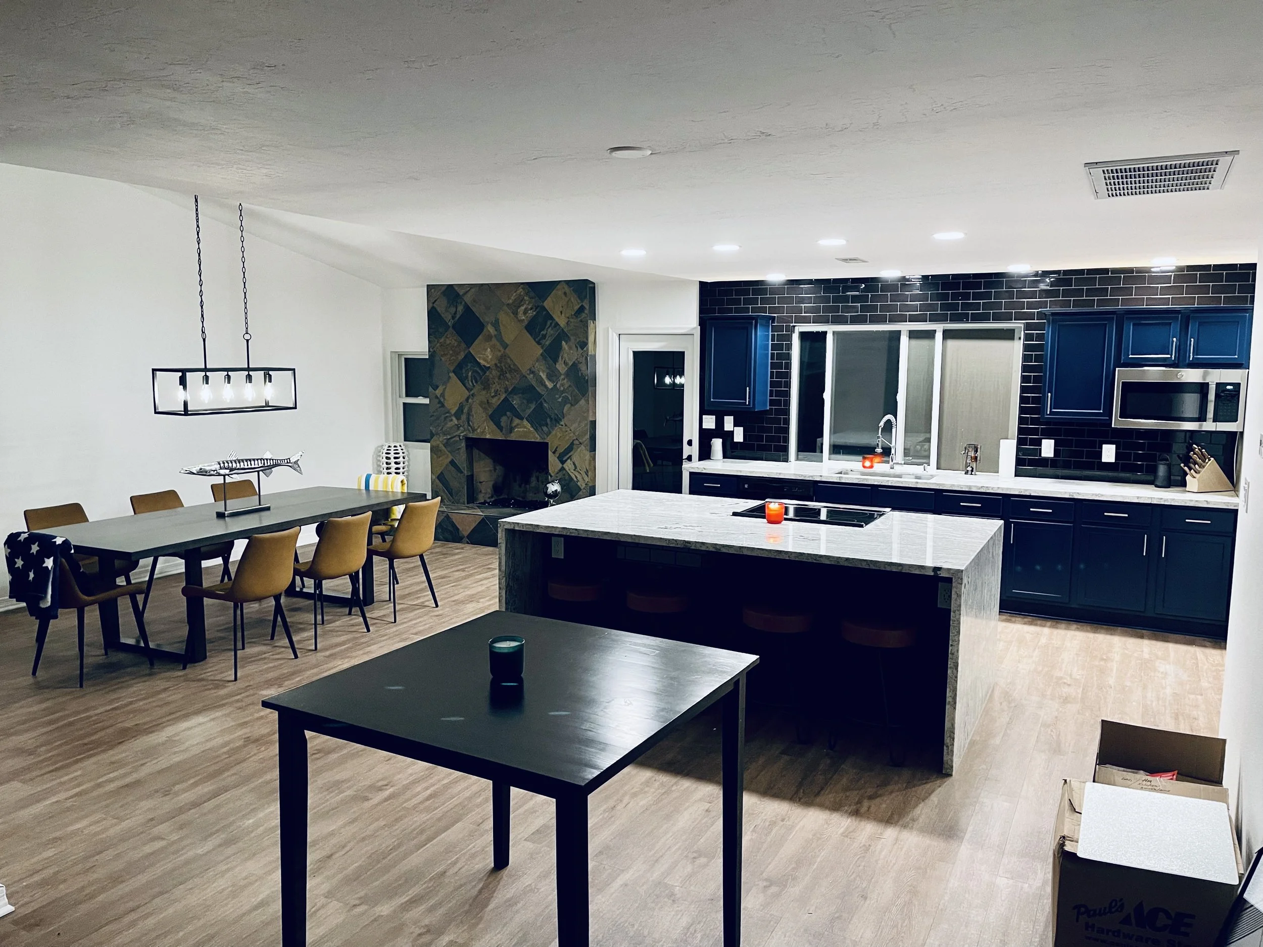 Modern kitchen and dining room with dark blue cabinets, a white marble island, black brick backsplash, a fireplace with a stone surround, and a dining table with yellow chairs.