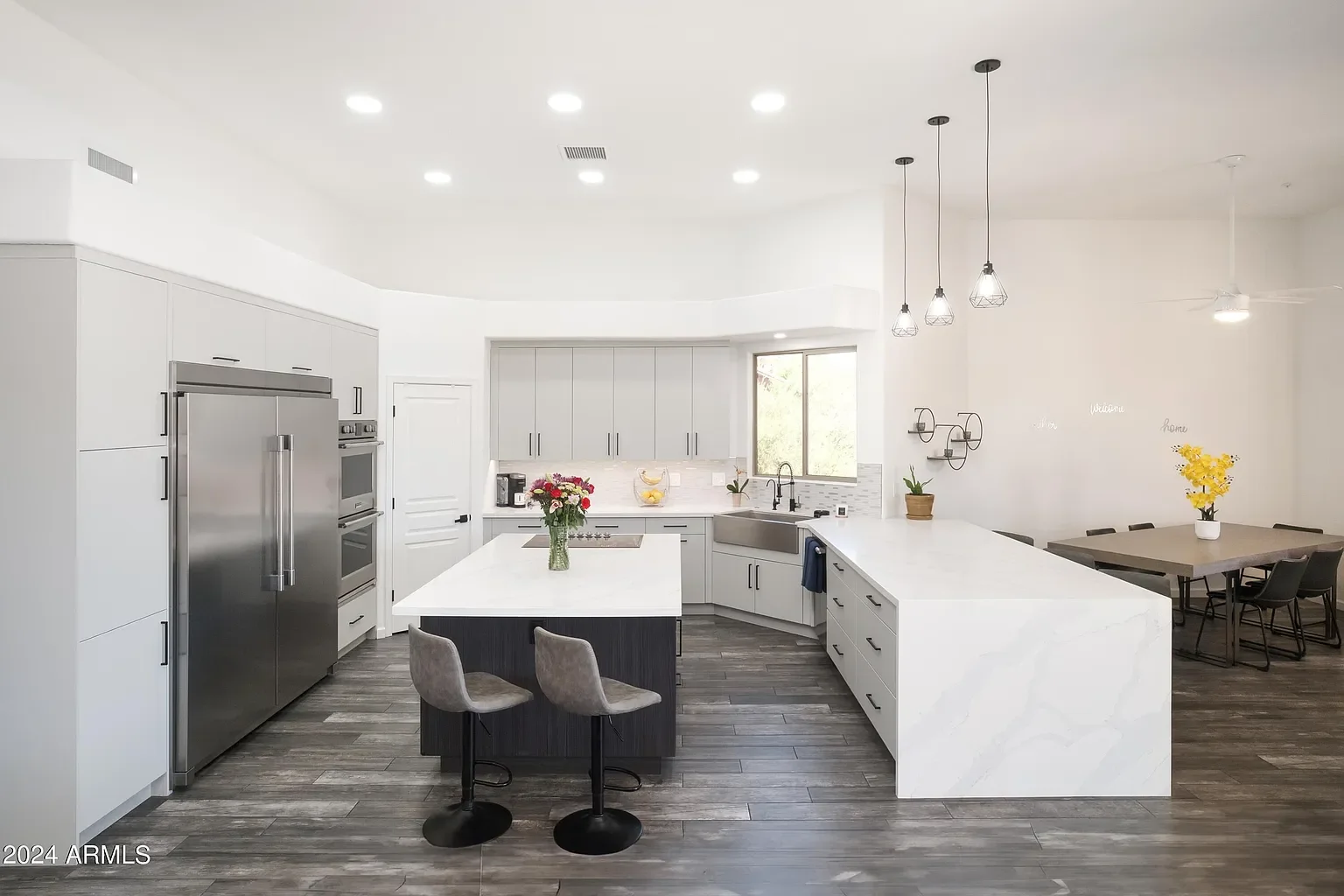 Modern kitchen with white cabinets, stainless steel refrigerator, island with flowers, dining area with table and chairs, pendant and ceiling lights, and hardwood floors.